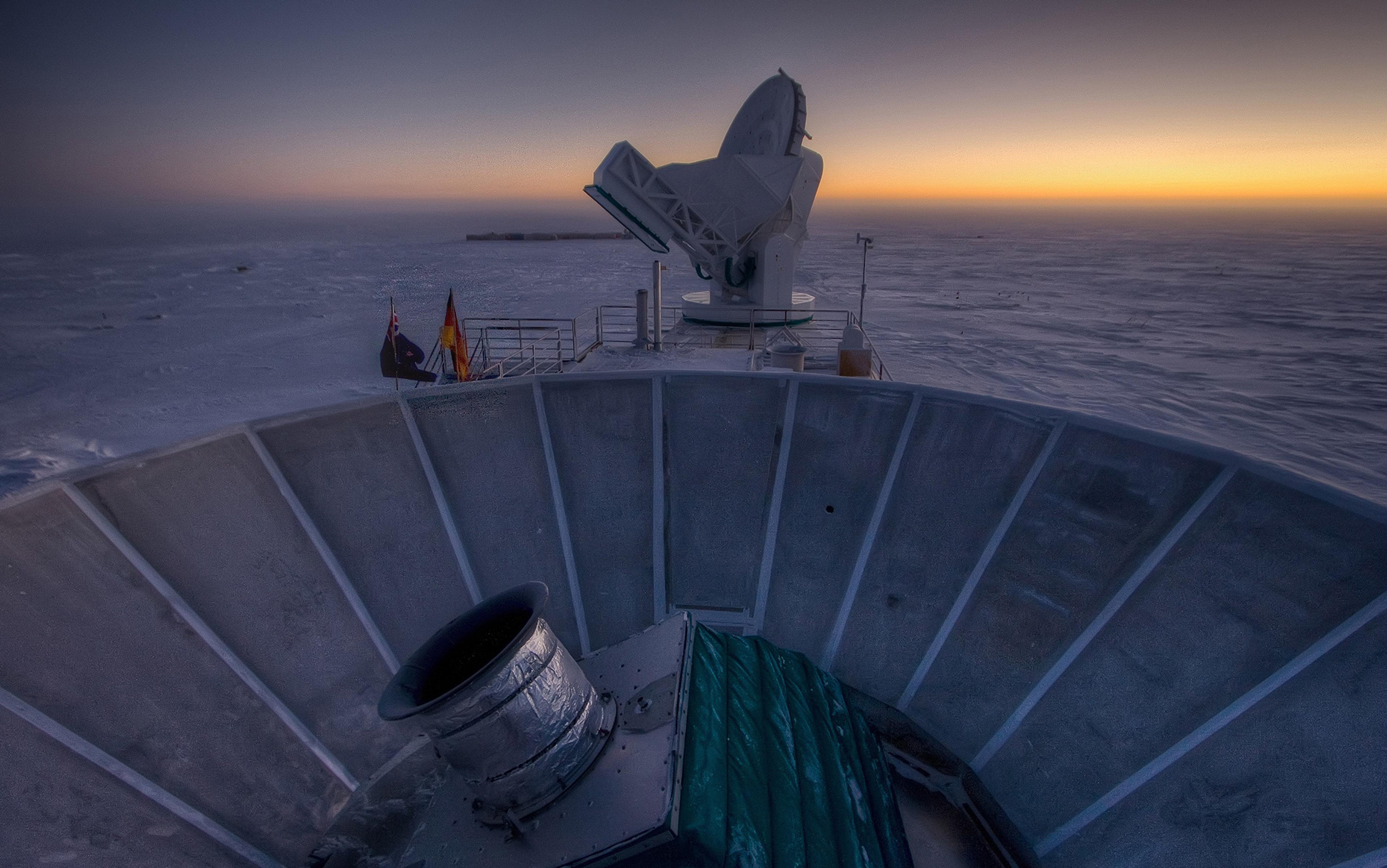 A snow-covered observatory at dusk with satellite dishes, flags and distant buildings, capturing an icy landscape and twilight sky.
