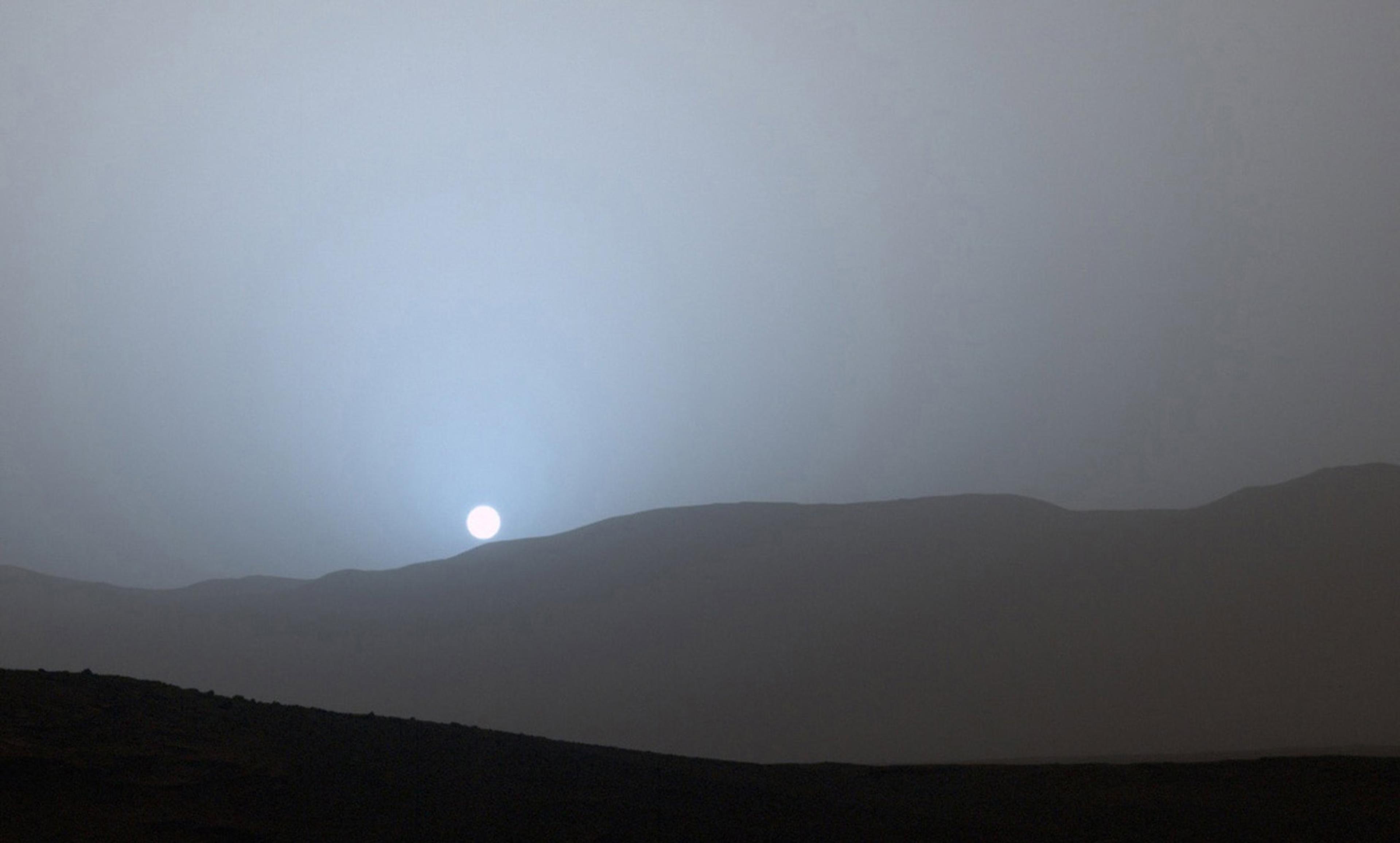 A pale sun setting behind a silhouette of rolling hills with a grey-blue sky, creating a serene, earthy vista.