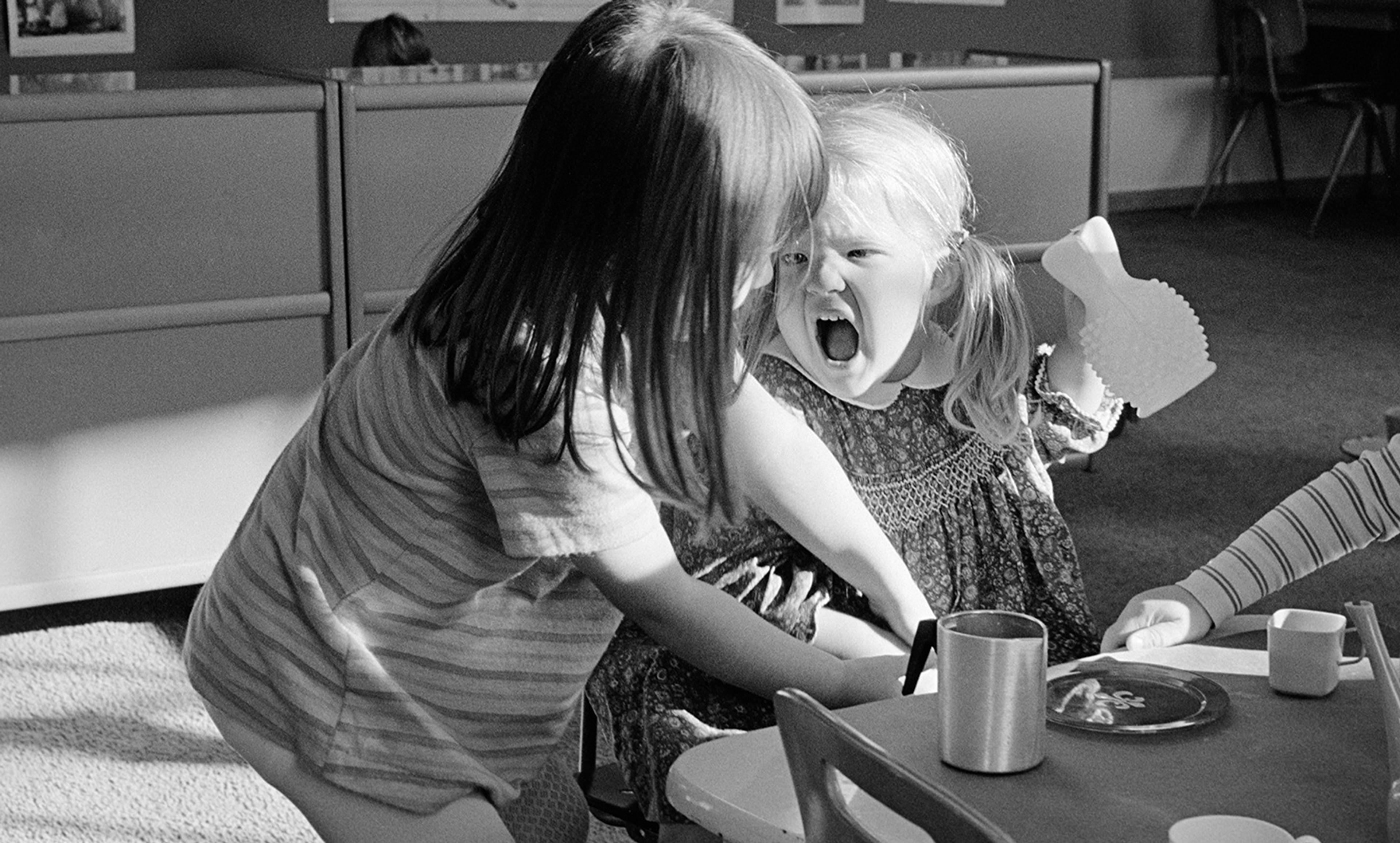 Black-and-white photo of two young girls indoors, one shouting angrily while holding a toy as the other looks down.