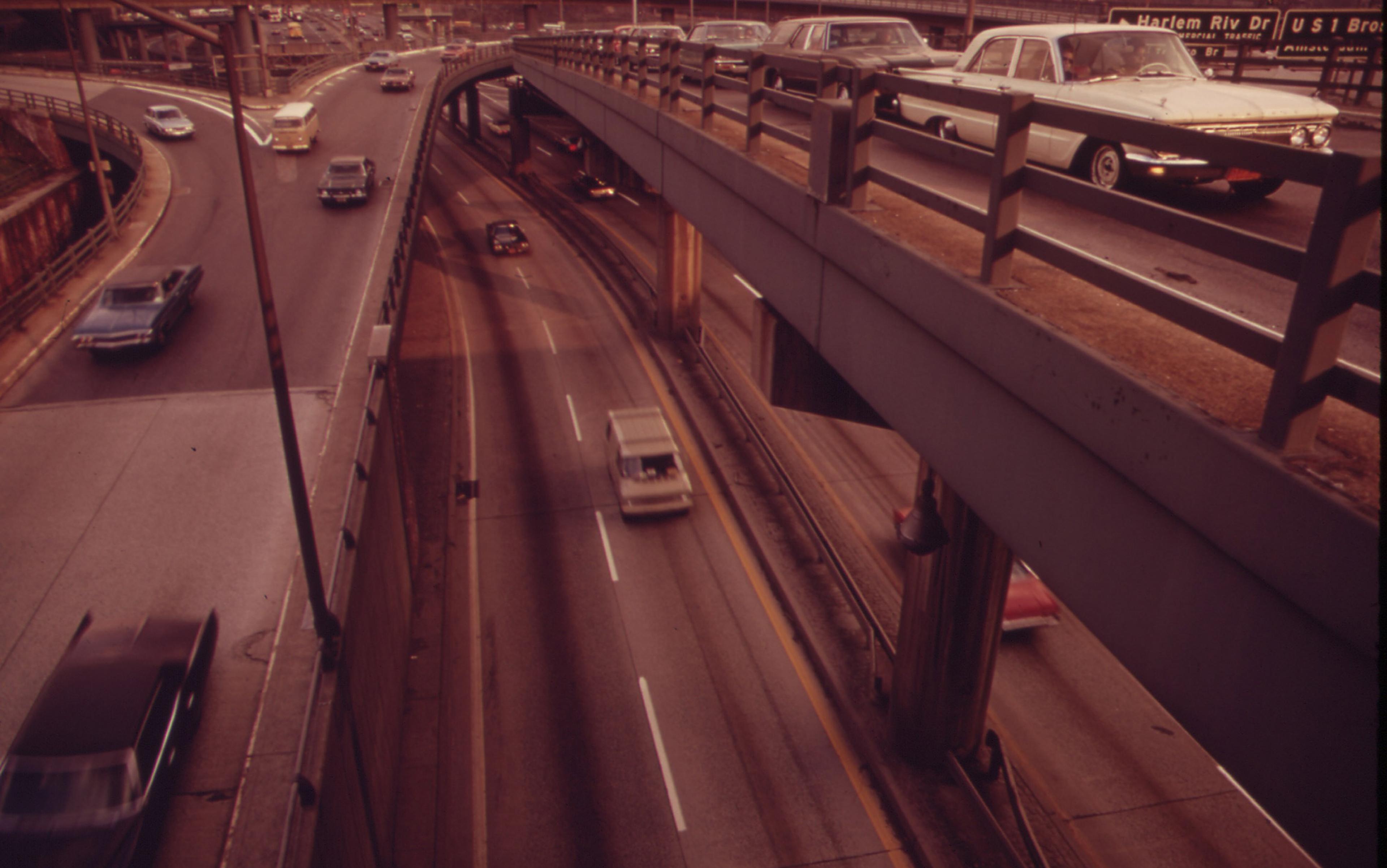 Aerial view of a multi-level motorway with older classic cars driving on both upper and lower levels, and infrastructure such as railings and signs visible.