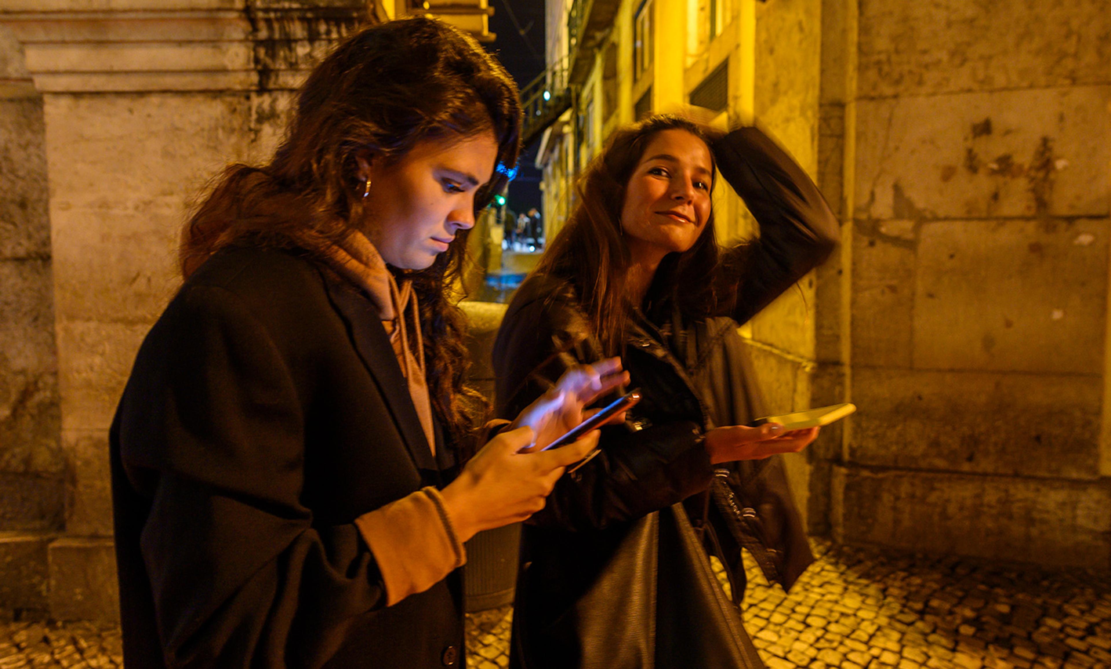 Two young women at night in a yellow-lit street, one looking at her phone, the other smiling and holding her phone with one hand.