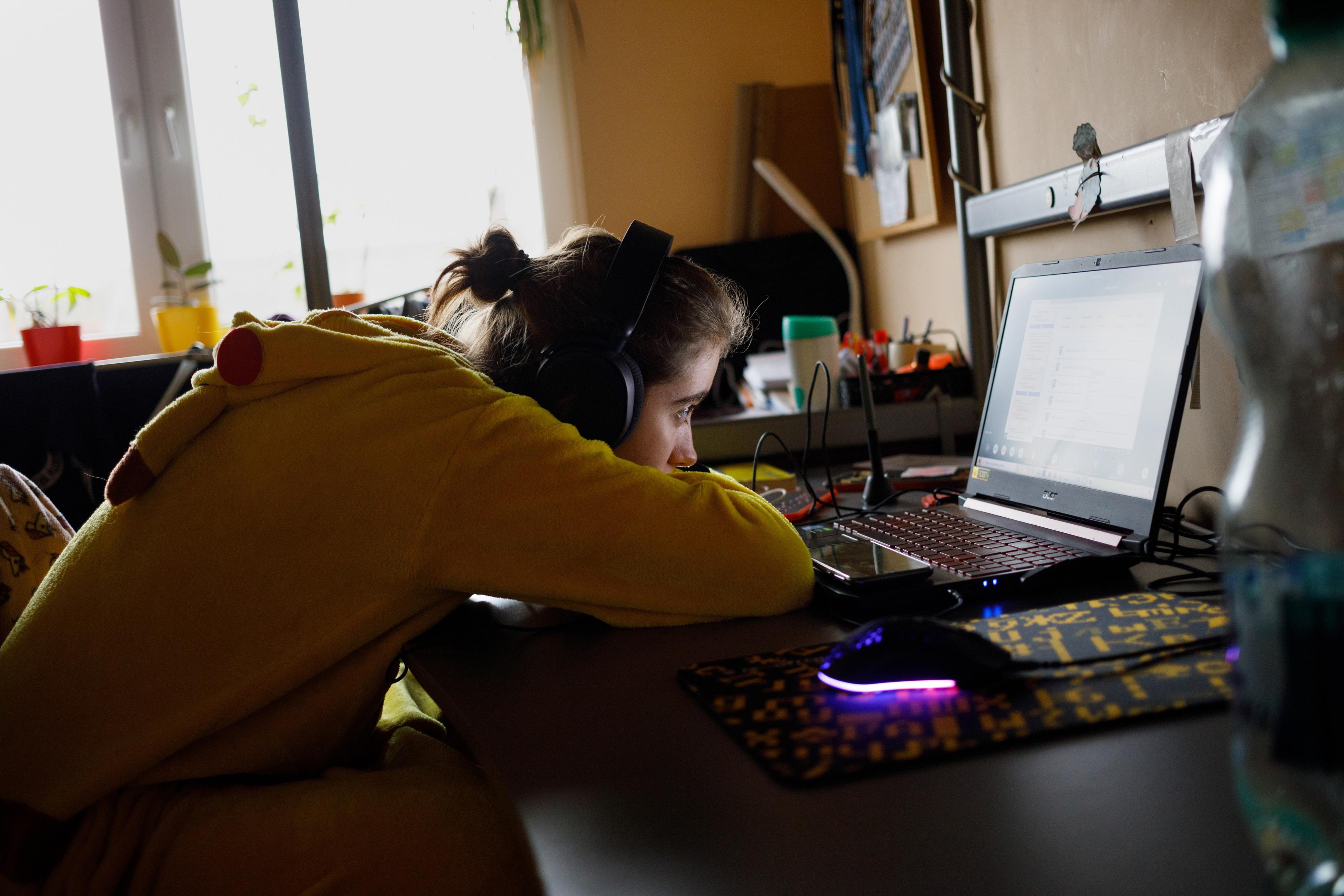 Photo of a person in a yellow onesie sitting at a desk using a laptop, with headphones on and a cluttered background.