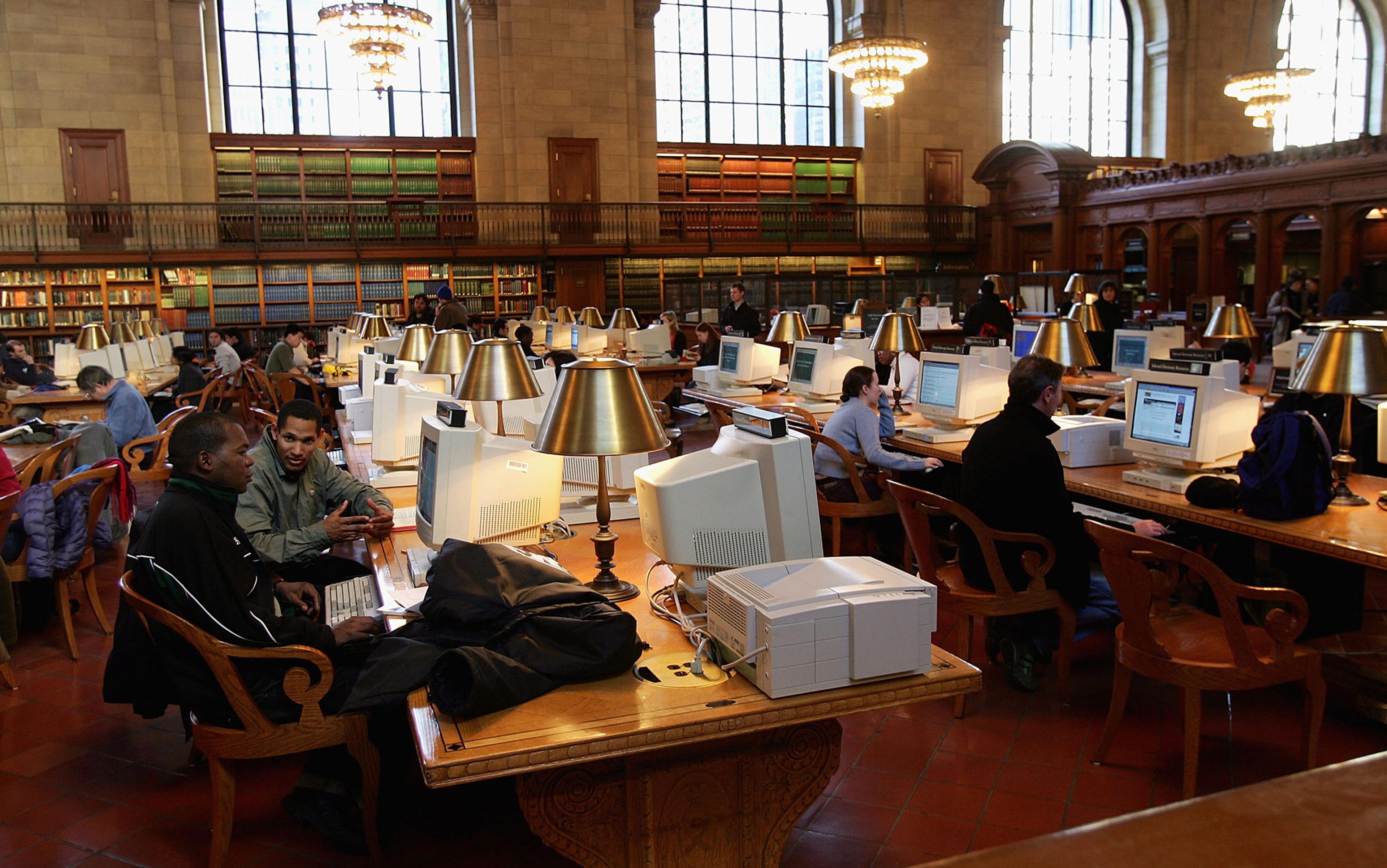 A busy public library reading room with people using desktop computers at wooden tables, surrounded by bookshelves and lit by large windows and chandeliers.
