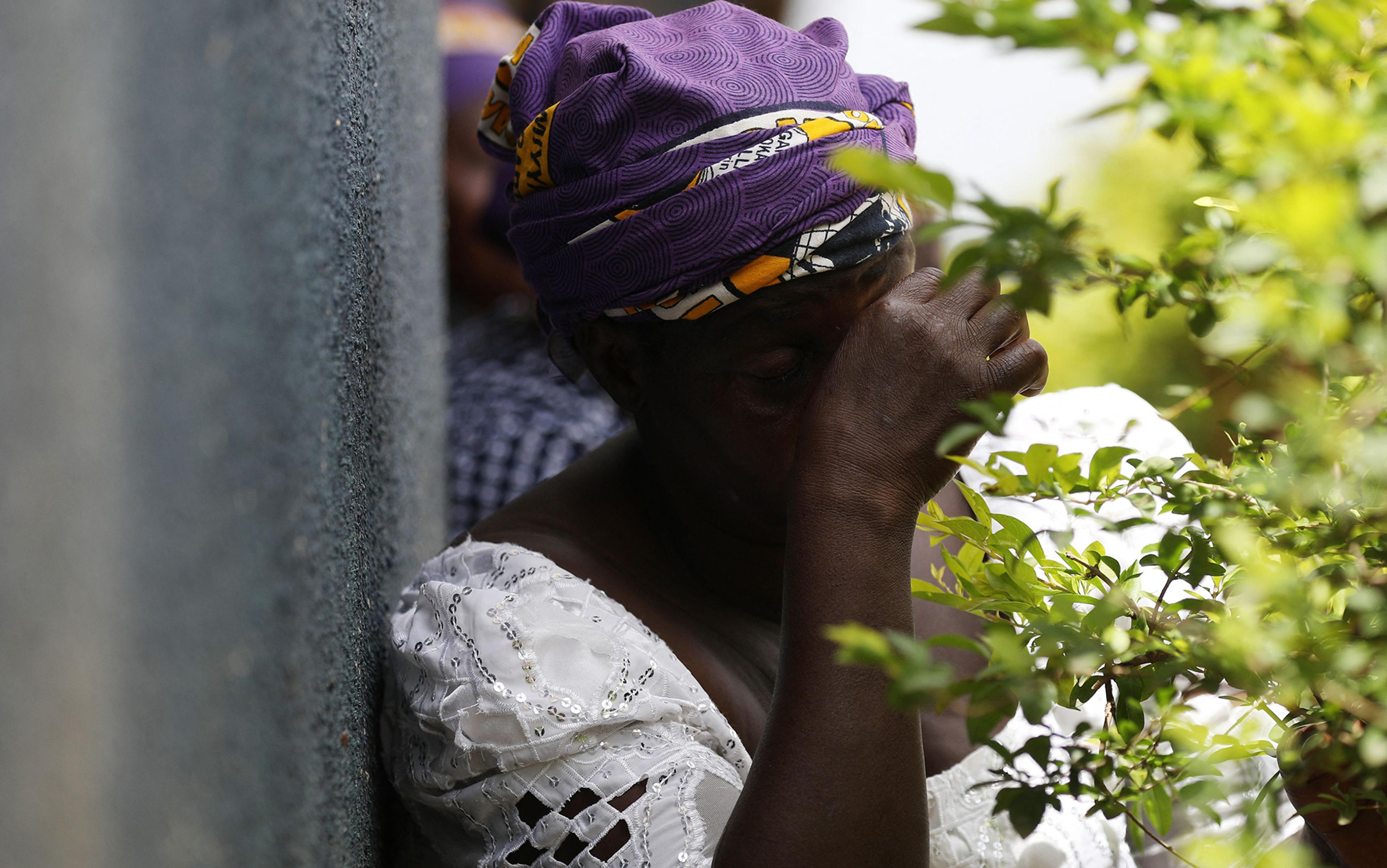 Photo of a woman in a white dress and purple headscarf leaning against a wall, covering her face near green foliage.