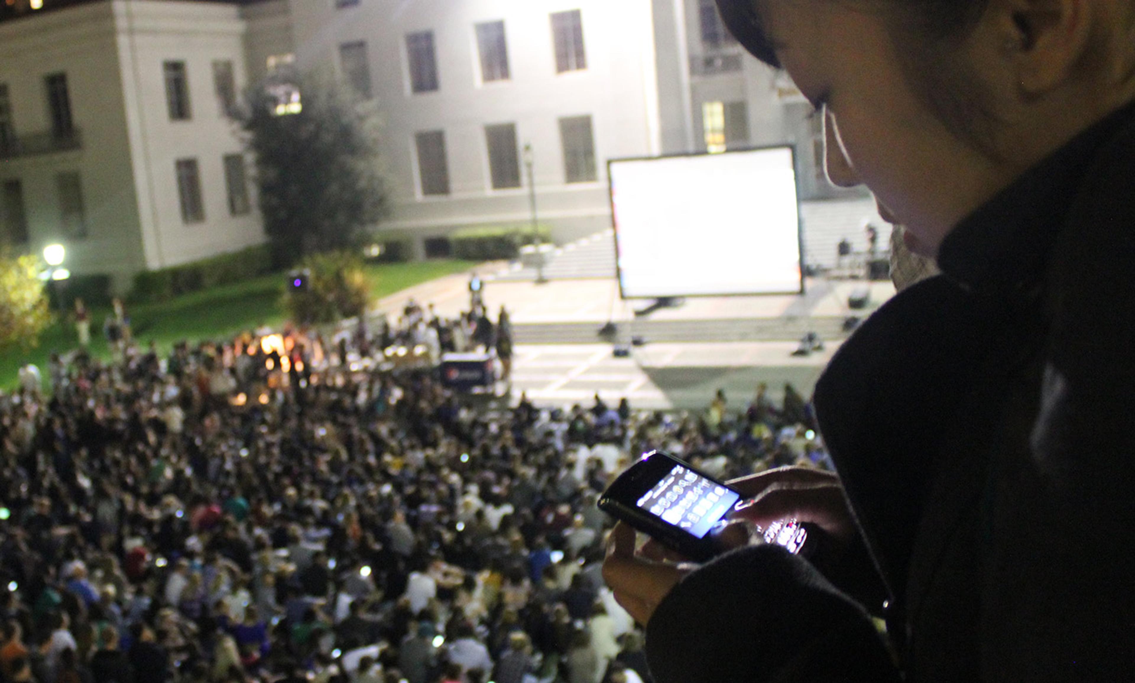 A person typing on a phone above a large crowd in front of a building with a projection screen at night.