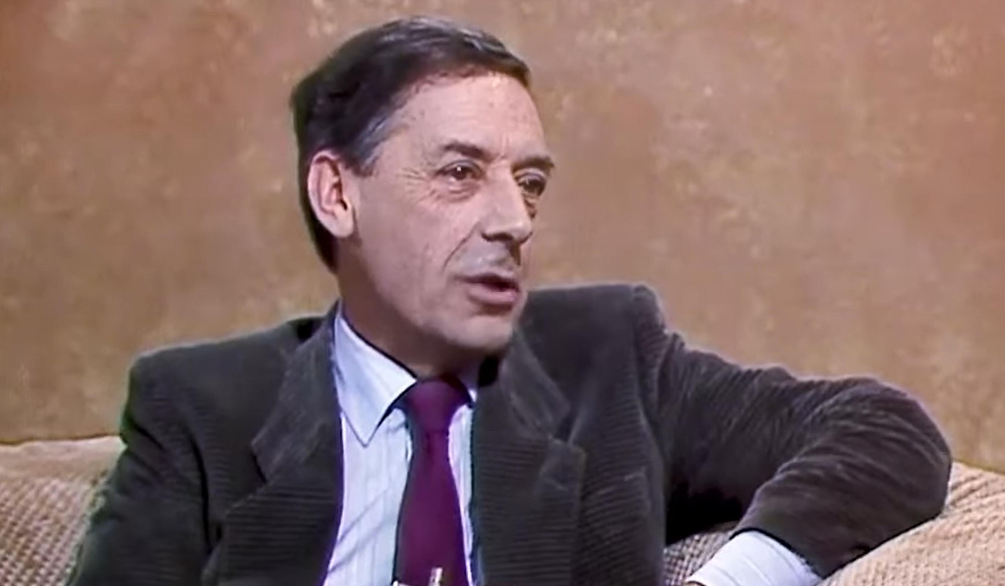 Man in a brown jacket and maroon tie sitting on a beige sofa, speaking with a neutral expression. Background is a plain, light brown wall.