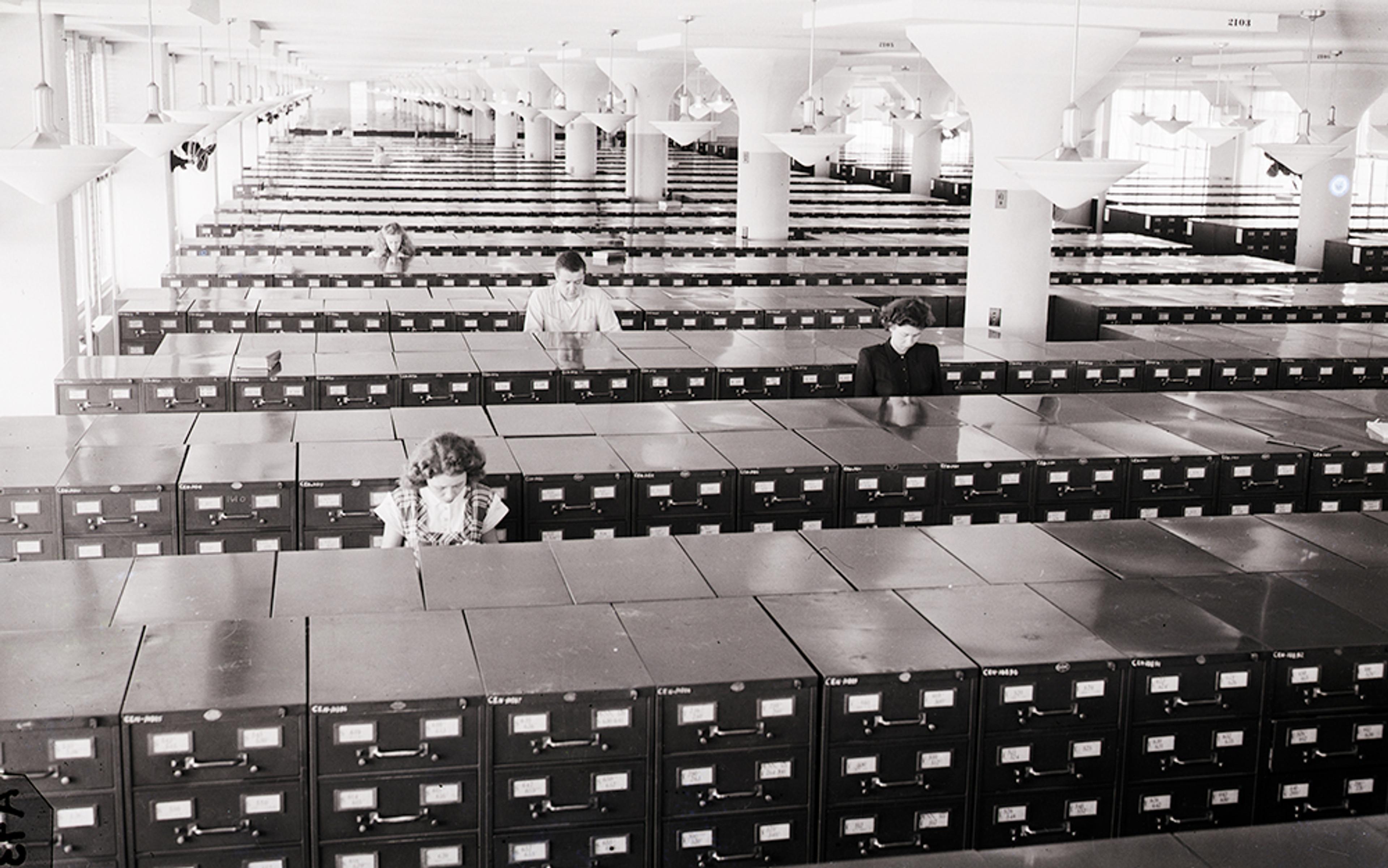 Black and white photo of people working in a large room filled with rows of filing cabinets in an office environment.