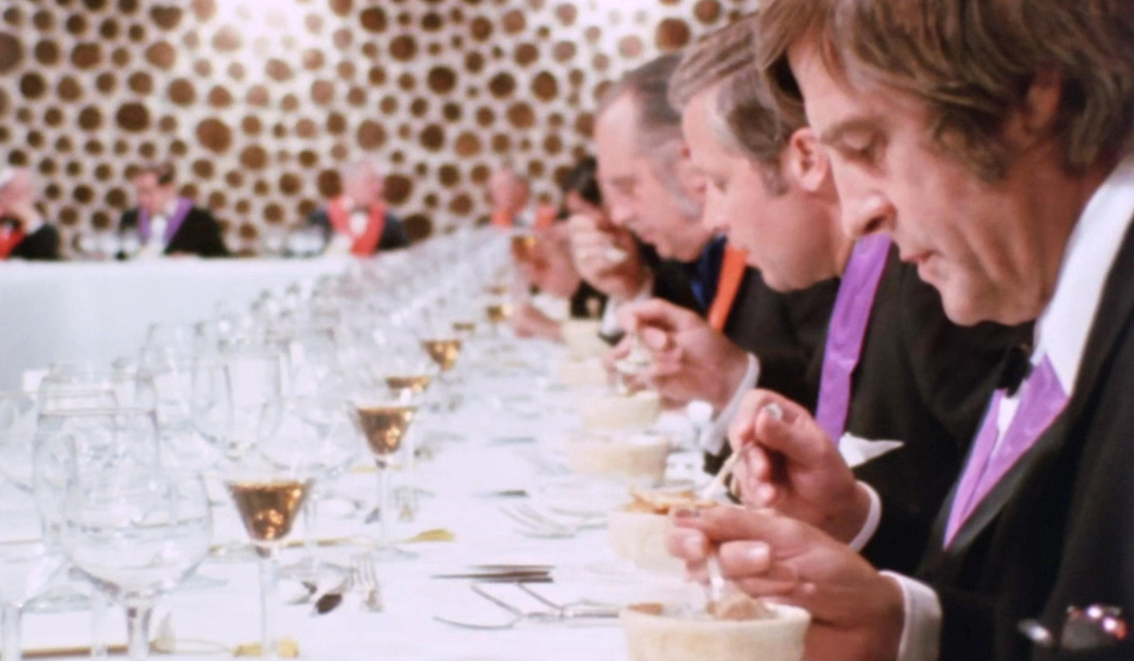 A group of men in formal attire sit at a long table, eating from bowls in a patterned room with numerous empty wine glasses.