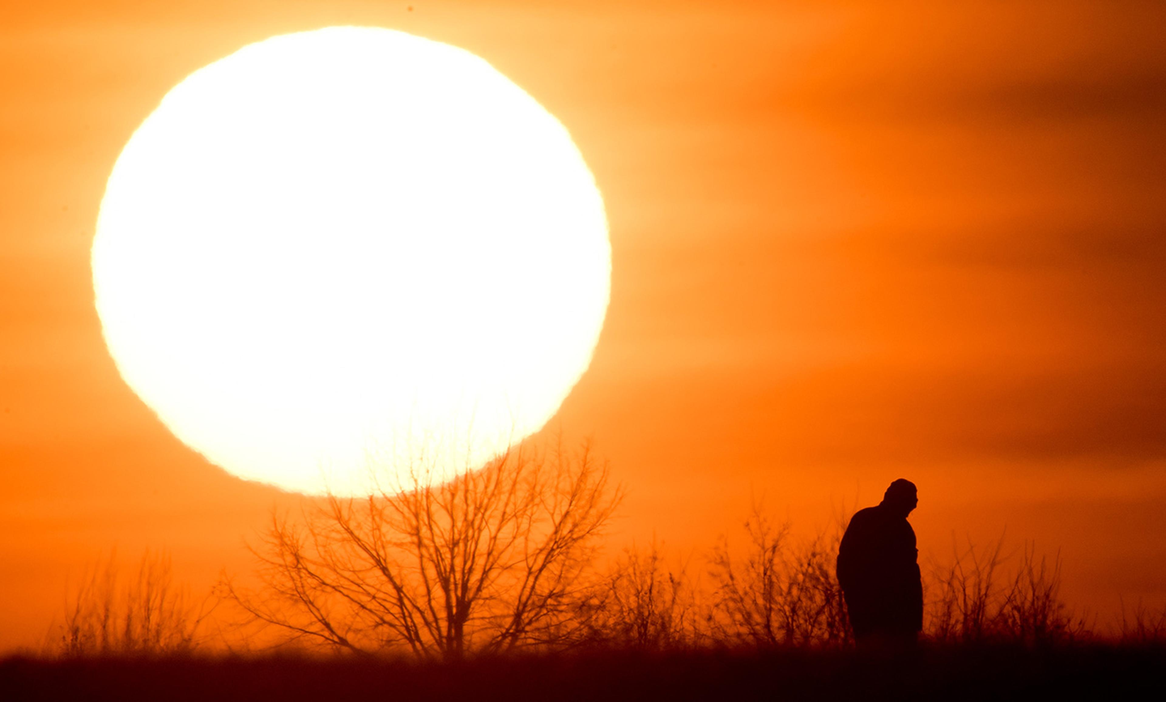 A silhouette of a person standing near trees with a large sun setting on the horizon, creating an orange sky background.