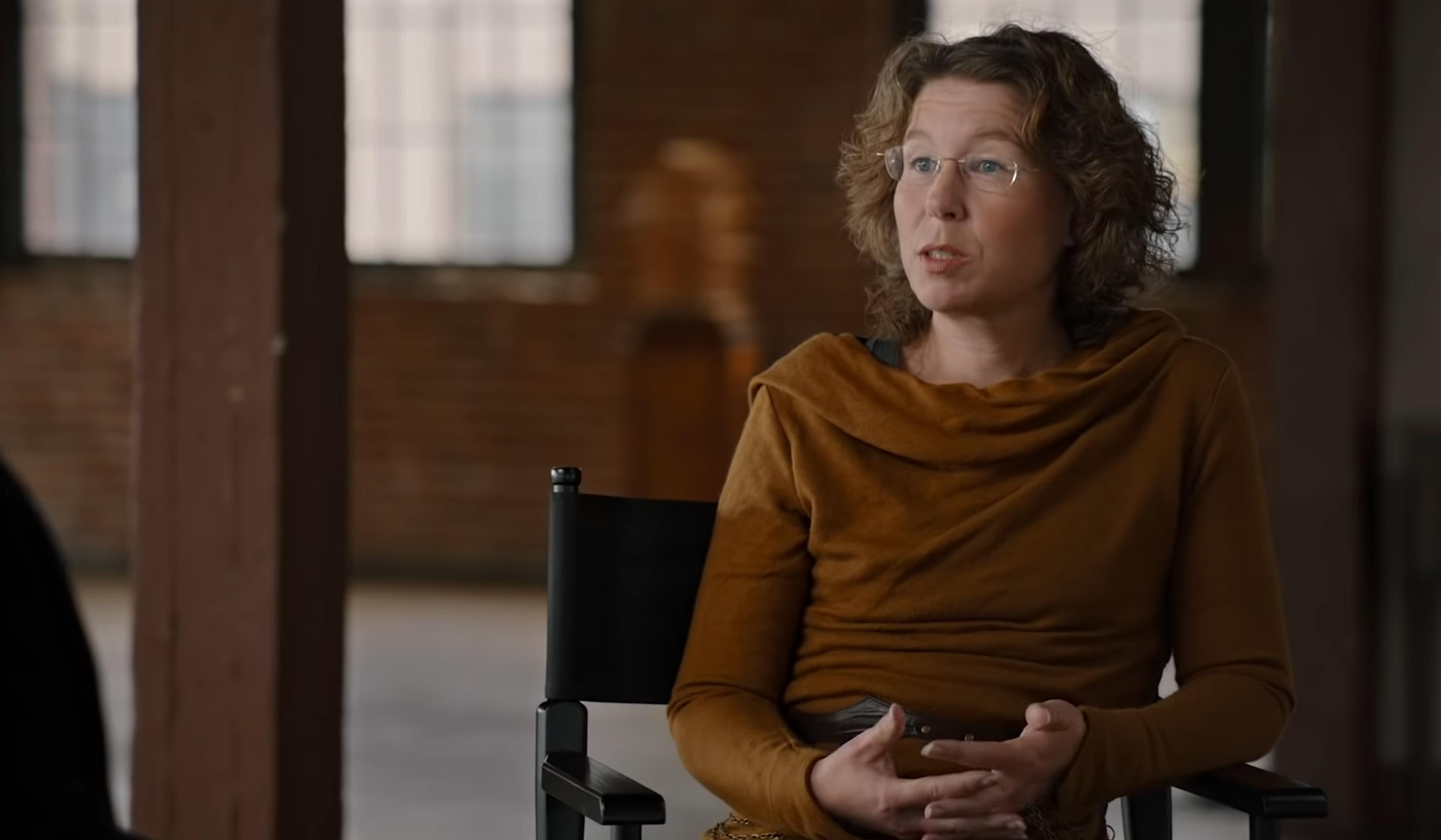 A woman with glasses and curly hair, wearing a brown top is speaking while sitting on a black chair.