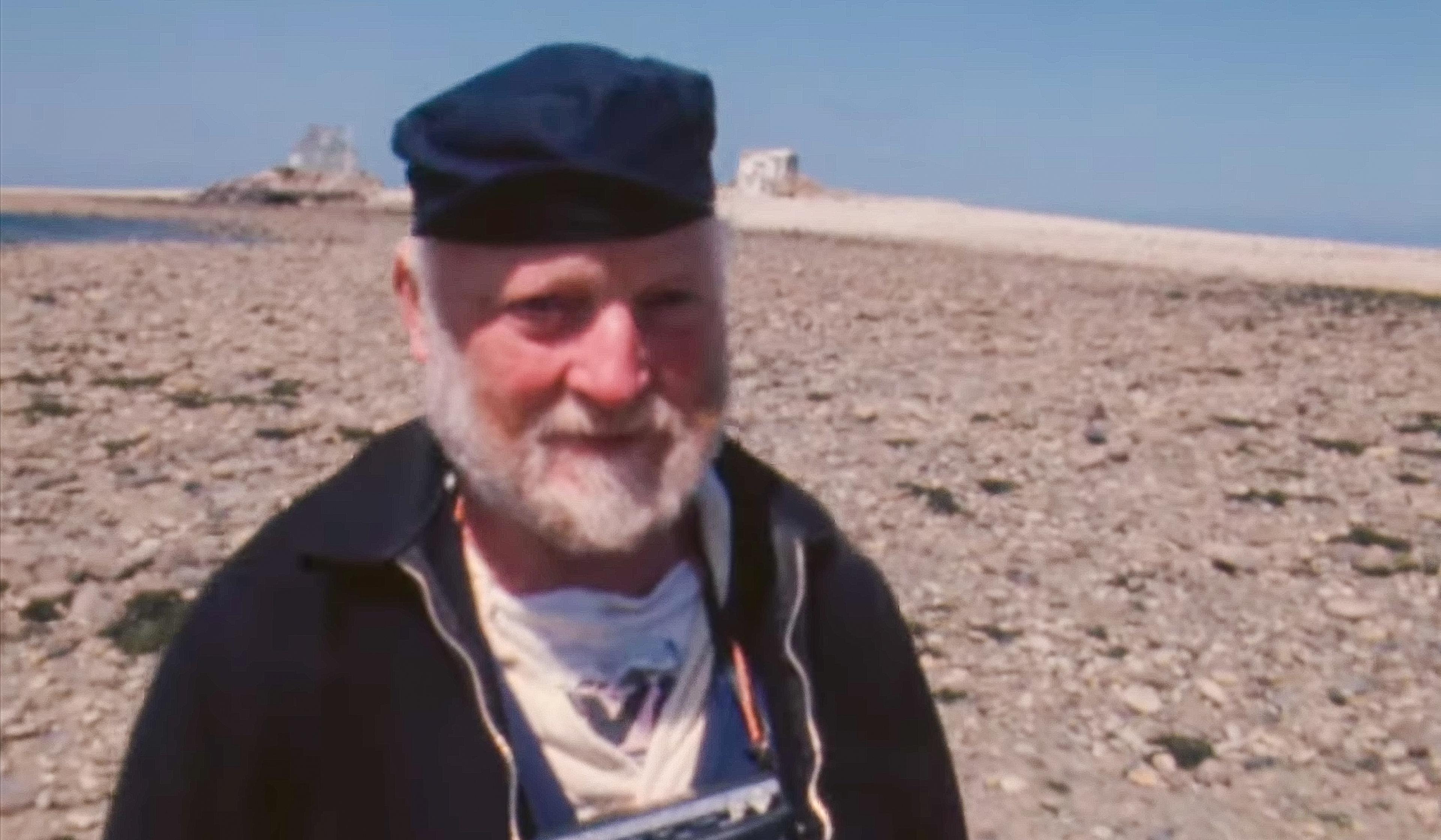 An elderly man with a white beard and blue cap stands on a rocky beach with ruins in the background on a clear day.