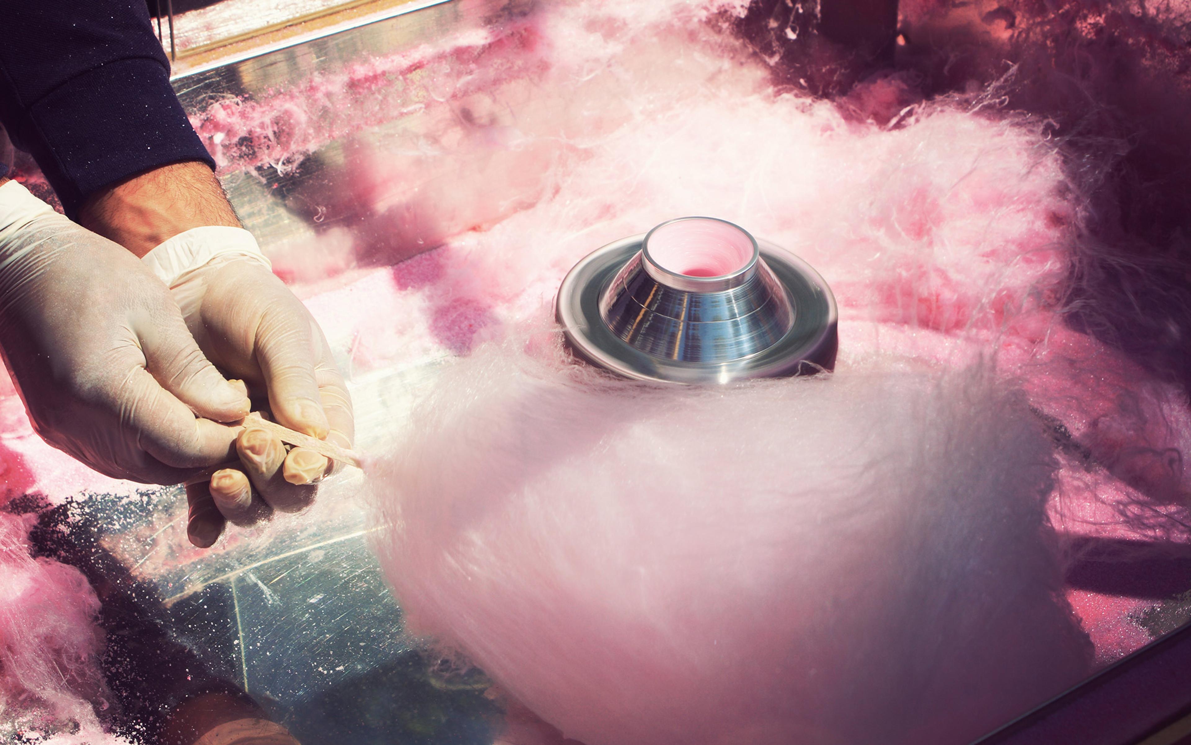 Hands wearing gloves making pink candyfloss with a machine, surrounded by fluffy pink candyfloss strands.
