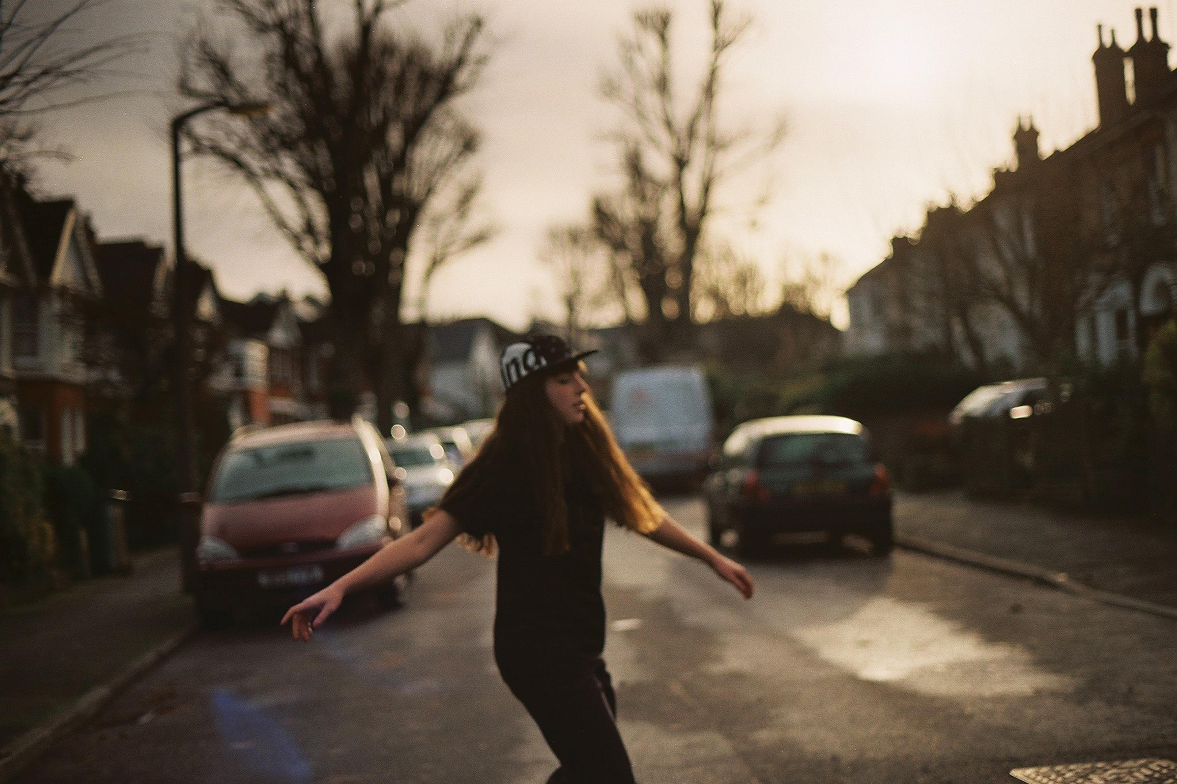 A person in a cap walking on a residential street with cars parked and bare trees in the background.