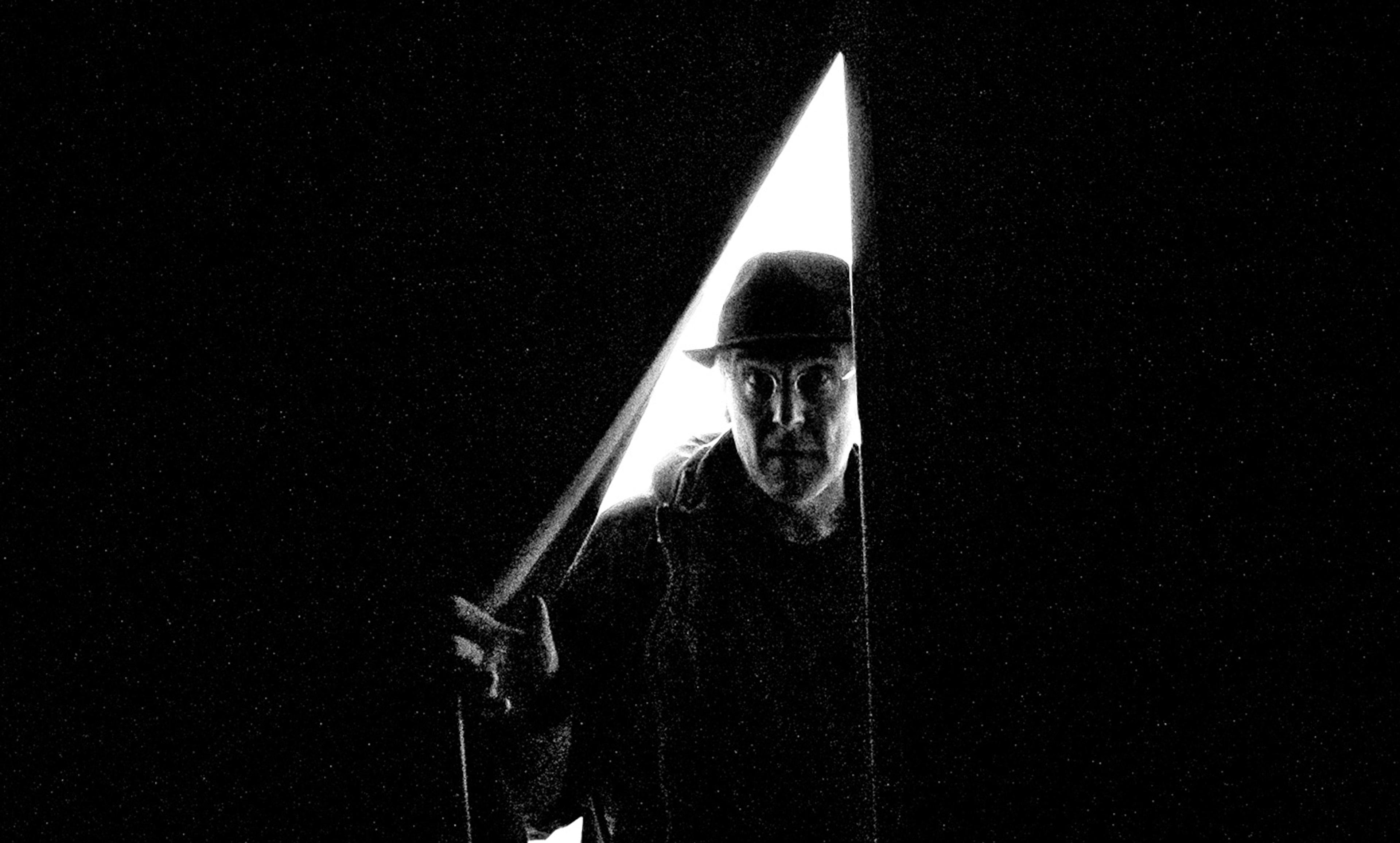 Black-and-white photo of a man in a hat partly revealed by a sliver of light through a door, creating a high-contrast, eerie scene.