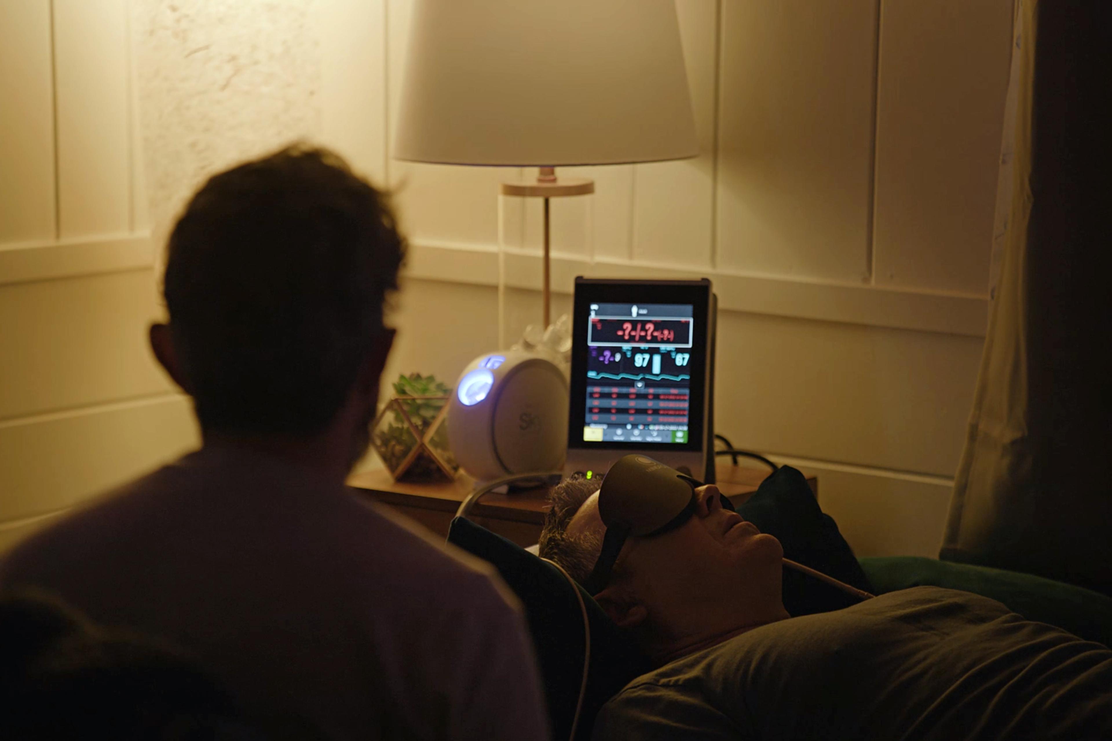 A man wearing an eye mask lies on a bed while another person observes; a monitor and lamp are on the nearby table.