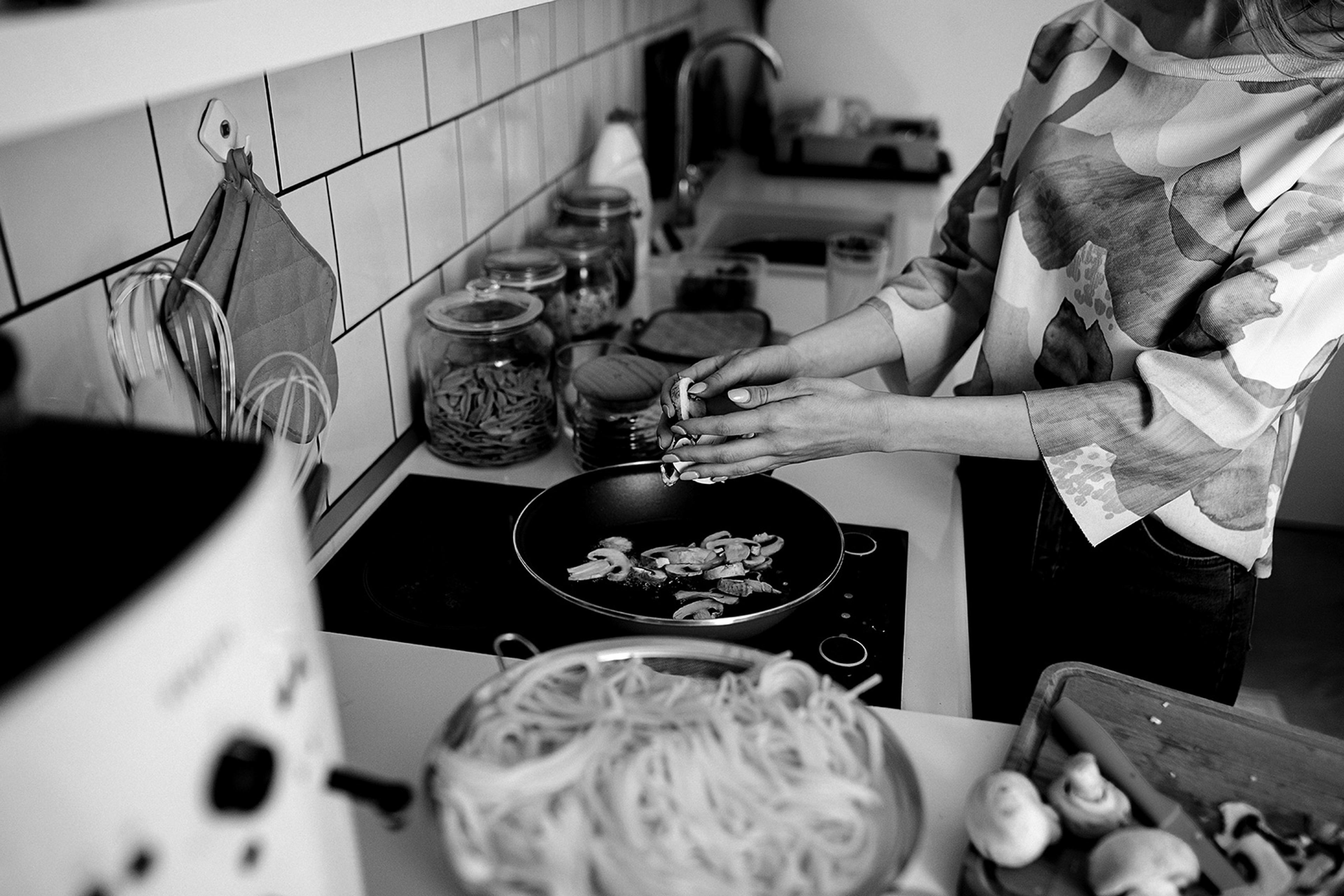 Black and white photo of a woman cooking in a kitchen with pasta and mushrooms on the counter.