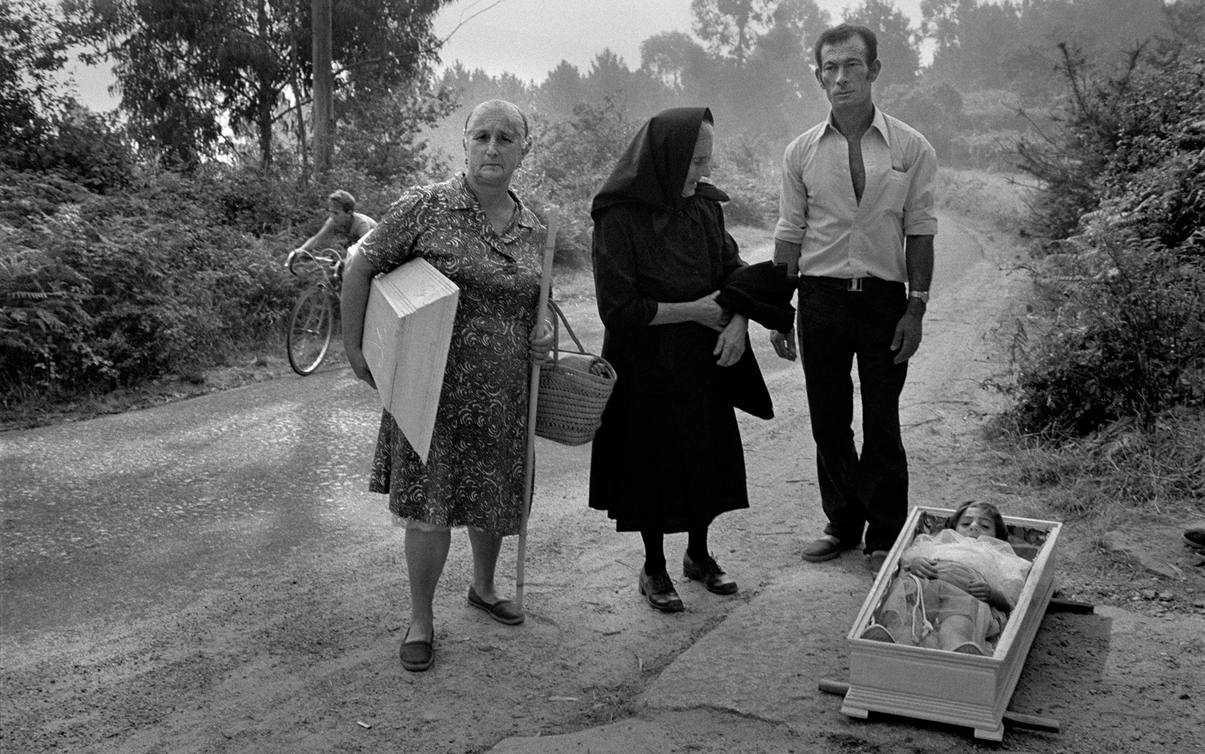Vintage photo of three adults next to a child in a coffin on a rural road, with a cyclist in the background among trees.