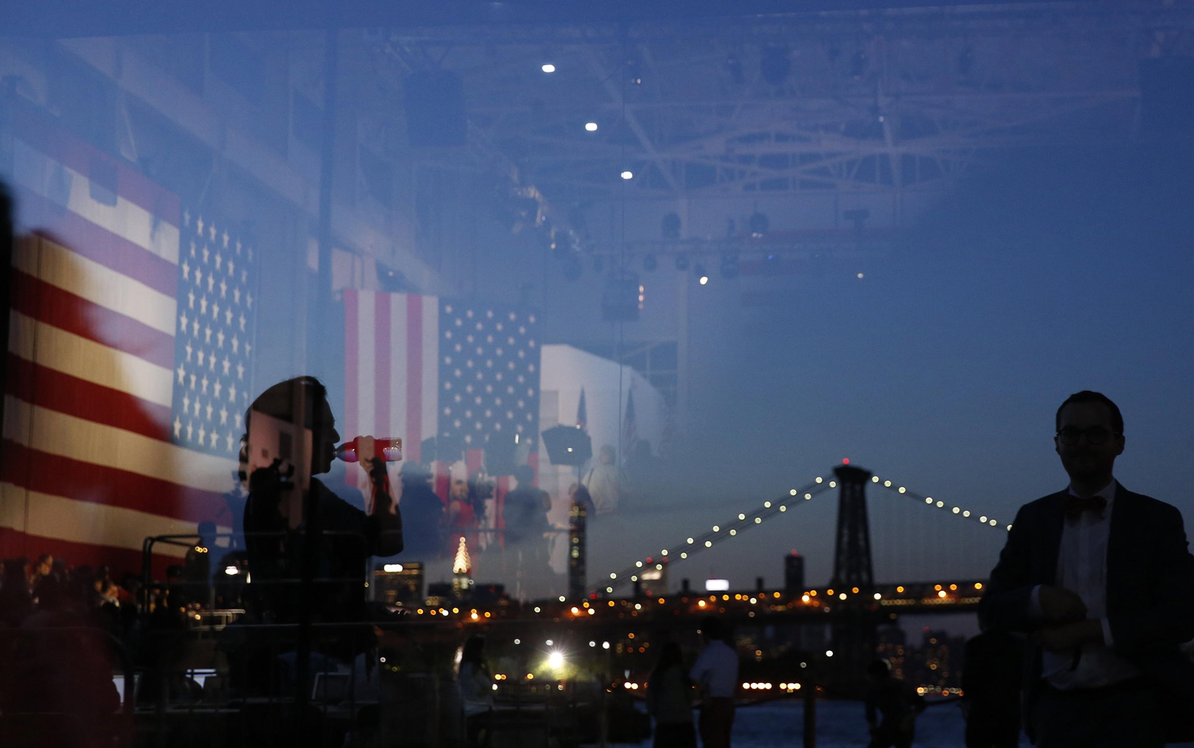 Photo showing the reflection of an indoor scene with American flags, silhouettes of people and an outdoor cityscape with a bridge at dusk.