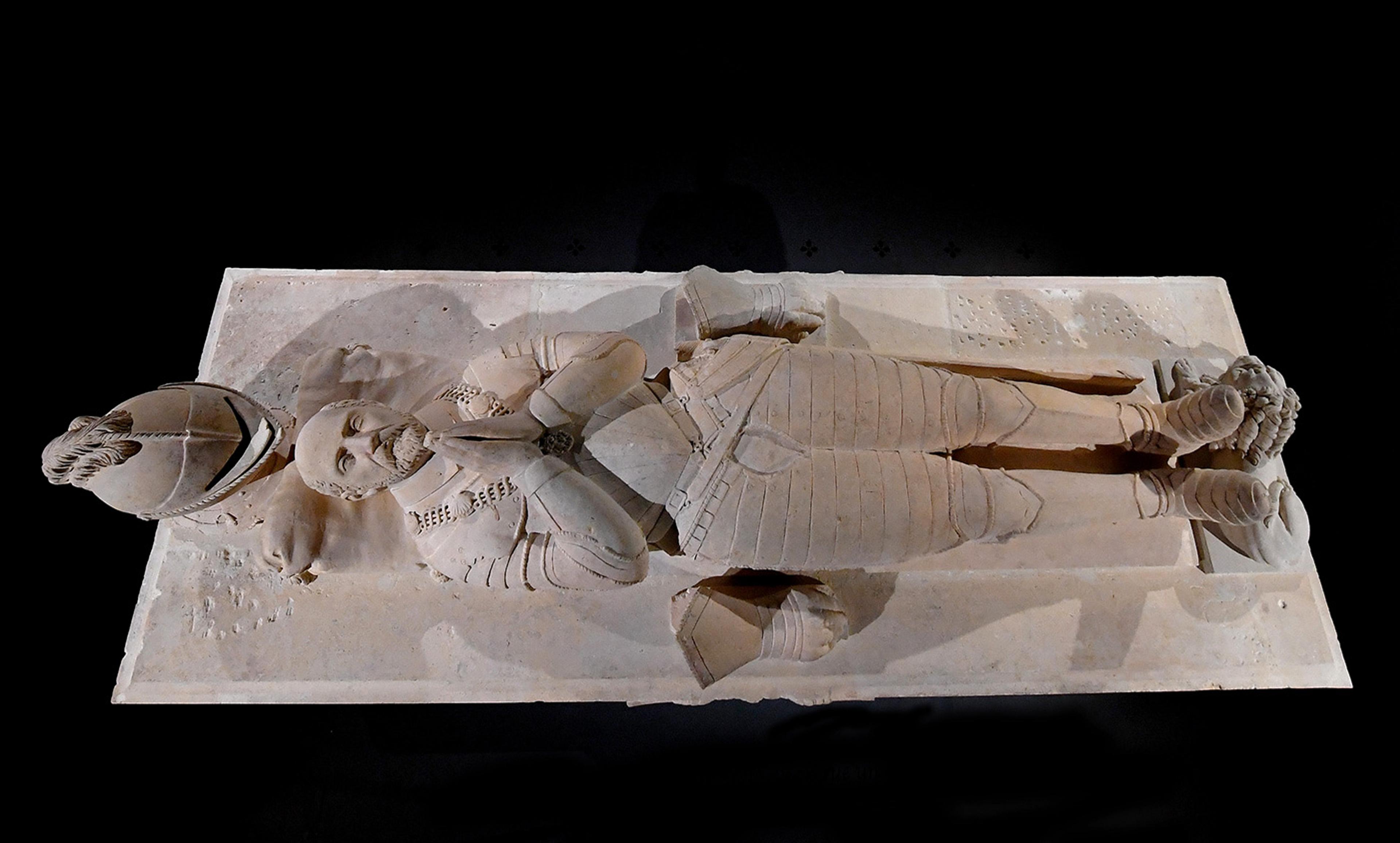 Stone effigy of a medieval knight in armour lying on a tomb with hands in prayer, a helmet beside him on a dark background.