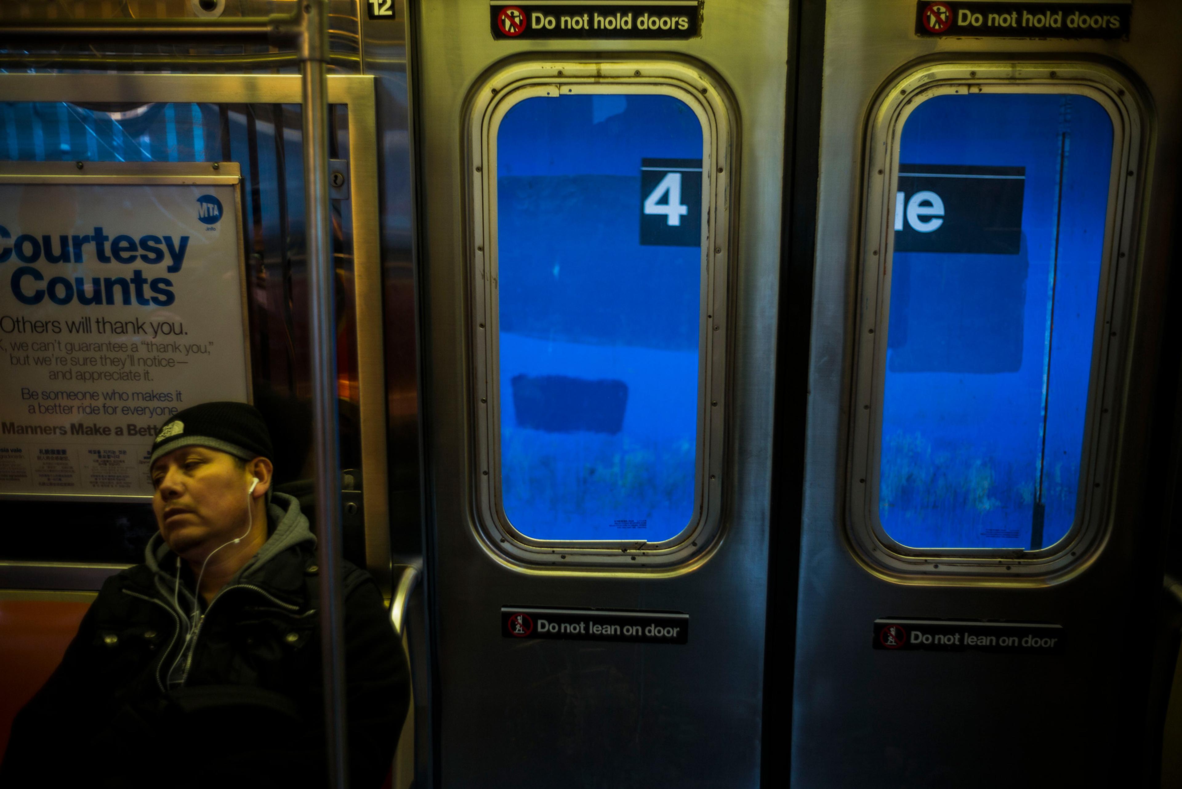 A person wearing headphones on a subway train next to a poster saying “Courtesy Counts”. Carriage doors have blue windows and “Do not hold doors” signs.