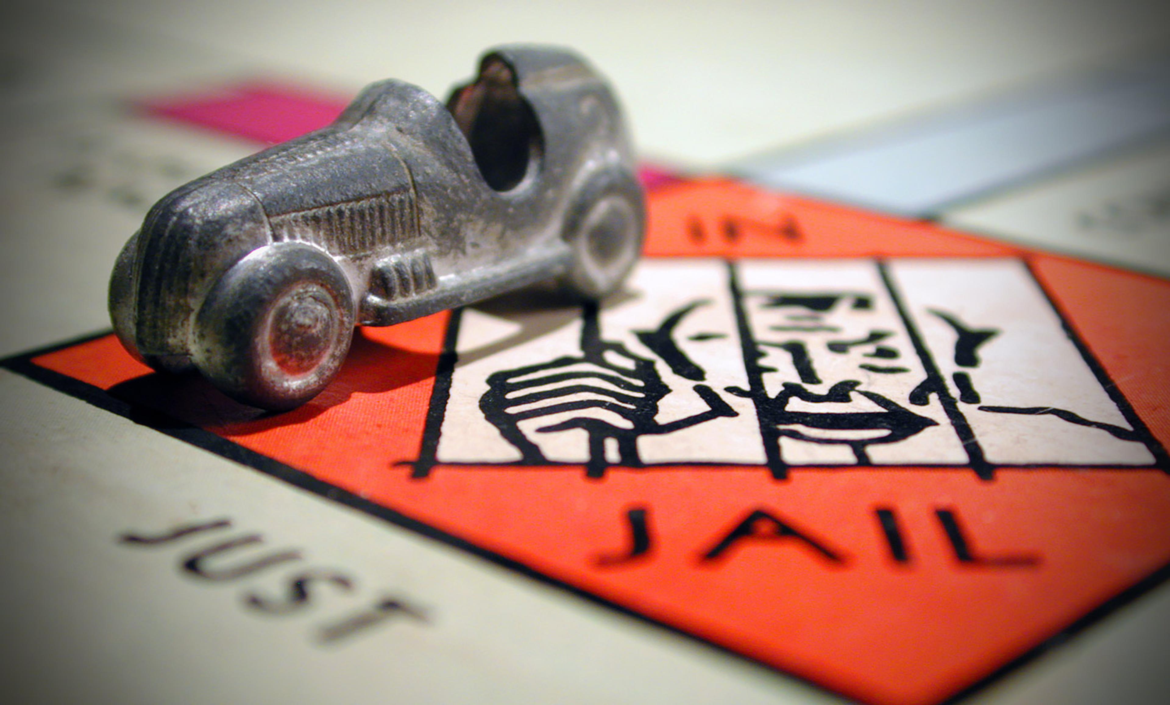 A metal car game piece on the “In Jail” square of a Monopoly board. The background shows blurred text and board squares.