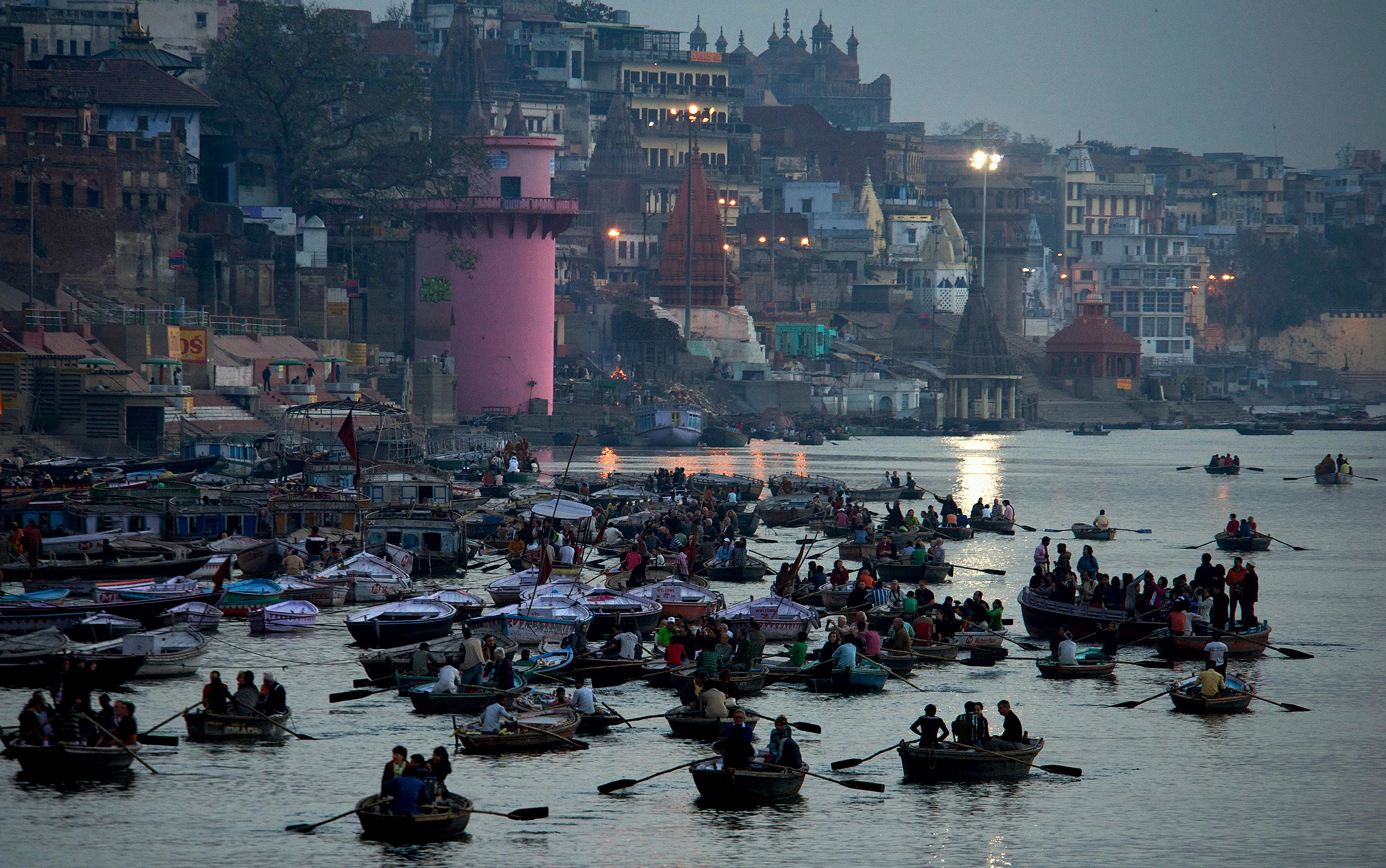 Dozens of small boats filled with people row along a busy river, with a densely built cityscape and a pink tower in the background.