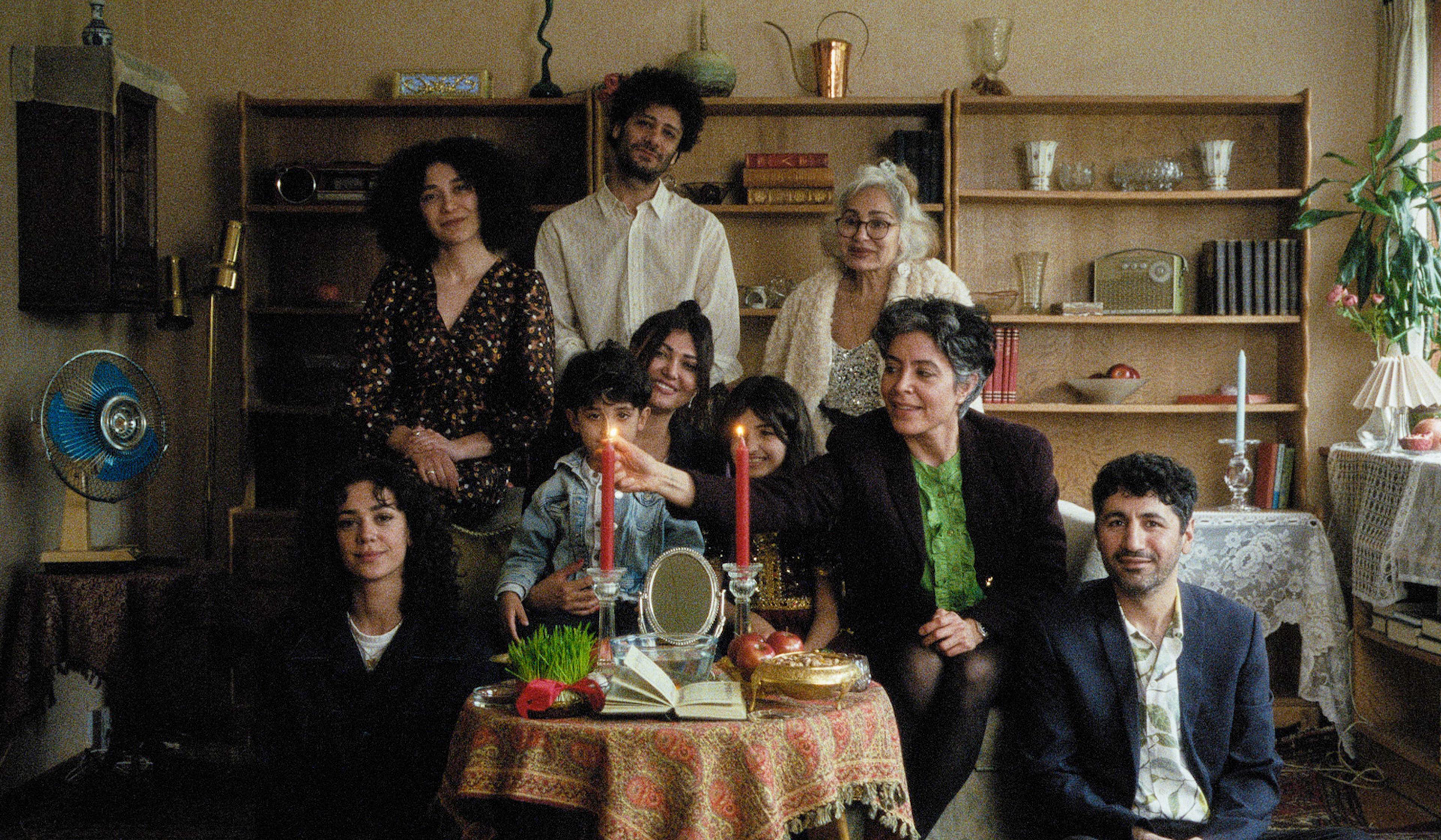 A smiling family group stands in a living room posing for a photo. The table is adorned with red candles, a mirror, apples, and a green plant.
