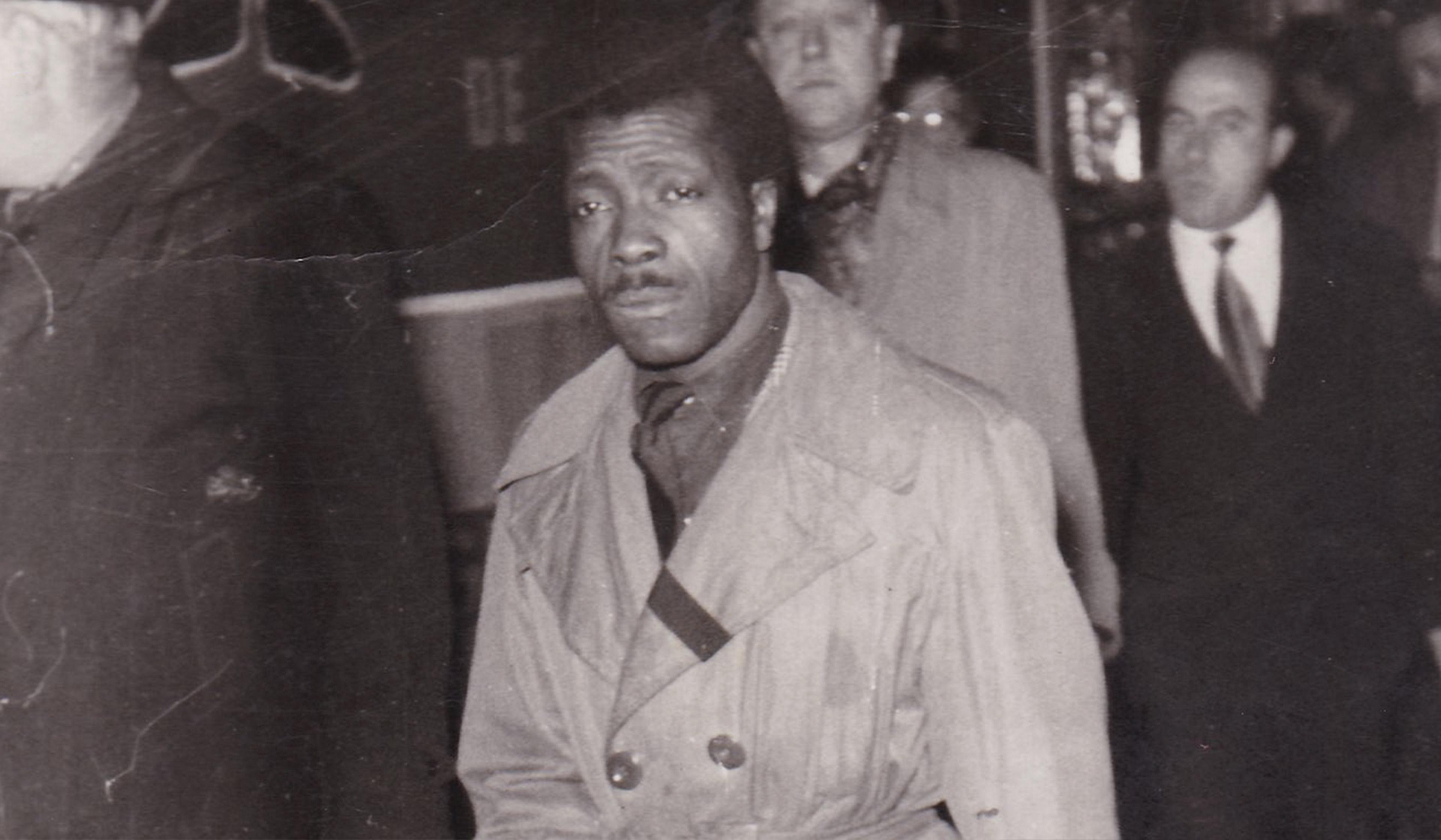 Black and white photo of a Black man in a trench coat walking, with a few other white men in suits visible in the background. He compellingly looks into the camera.