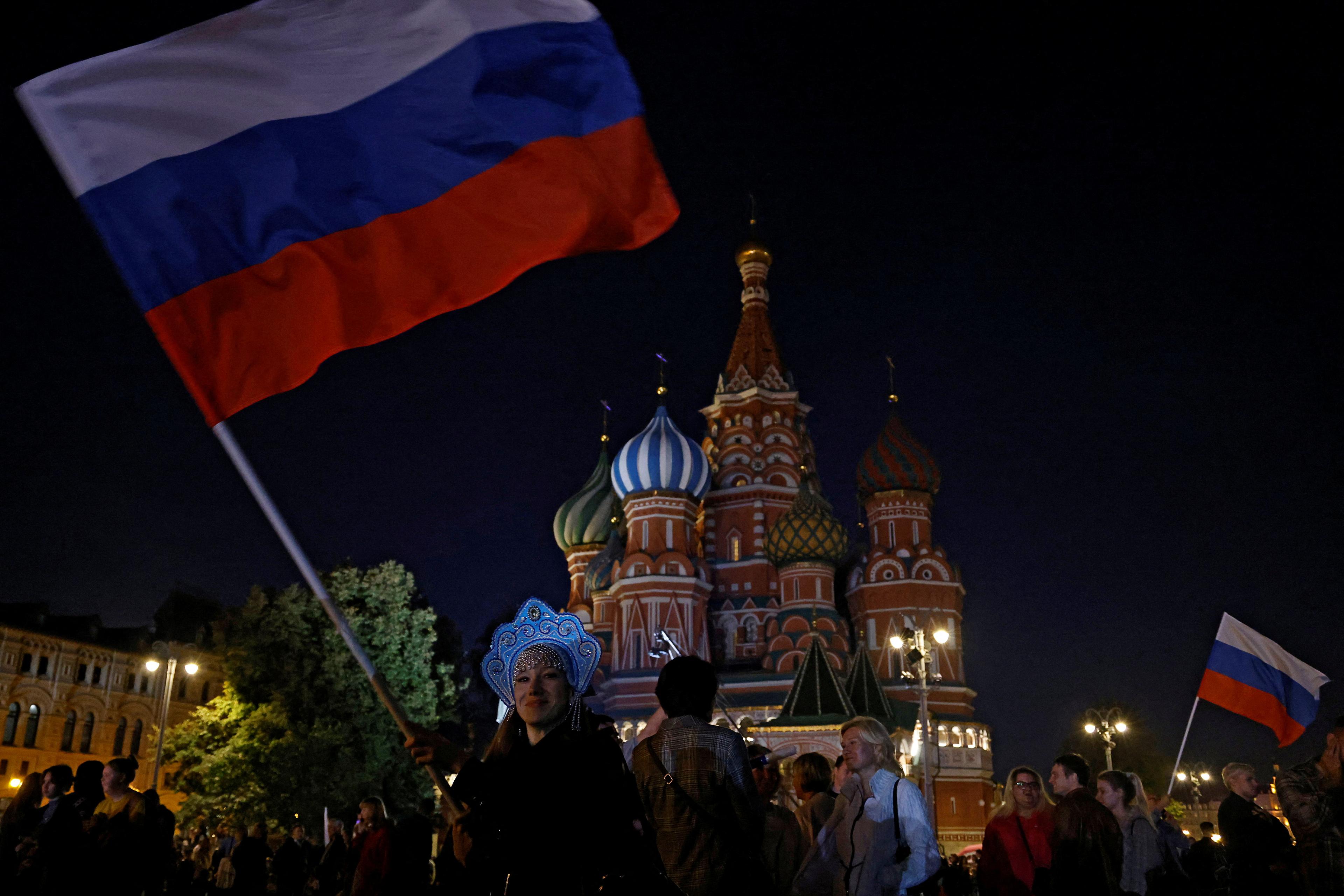 People at night in front of St Basil’s Cathedral in Moscow with a woman waving a large Russian flag in the foreground.