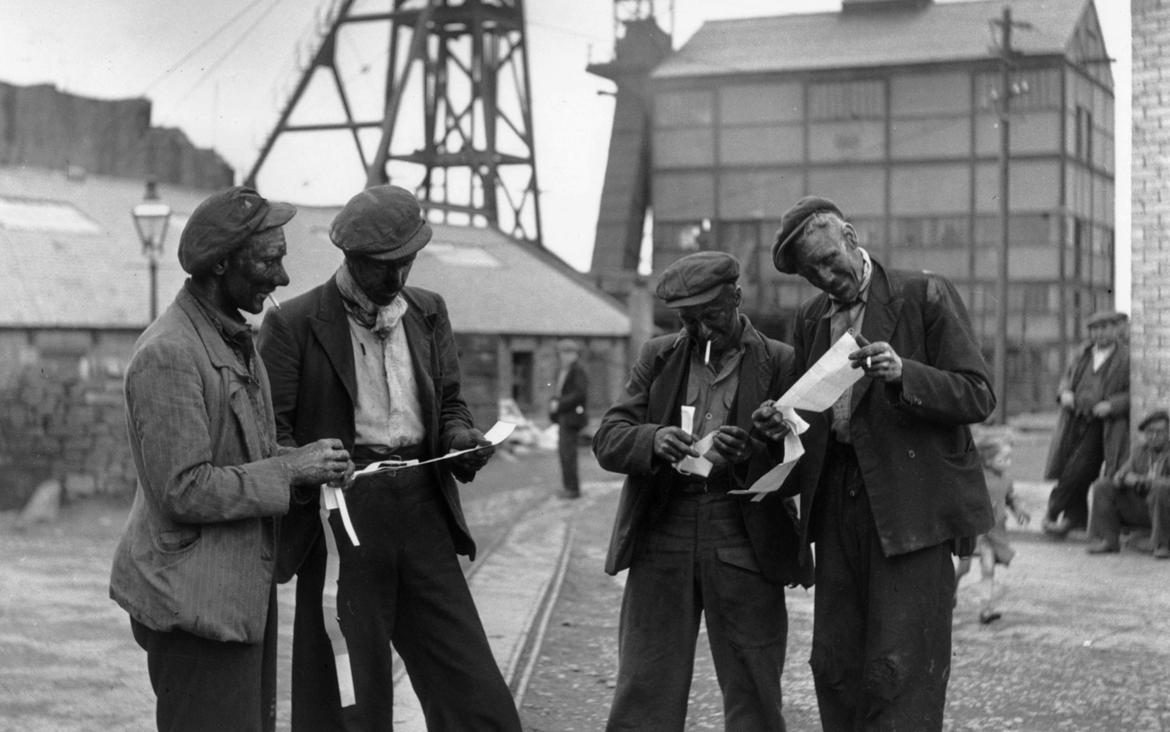 Black and white photo of four coal miners covered in dust examining paper slips outside an industrial building and machinery.
