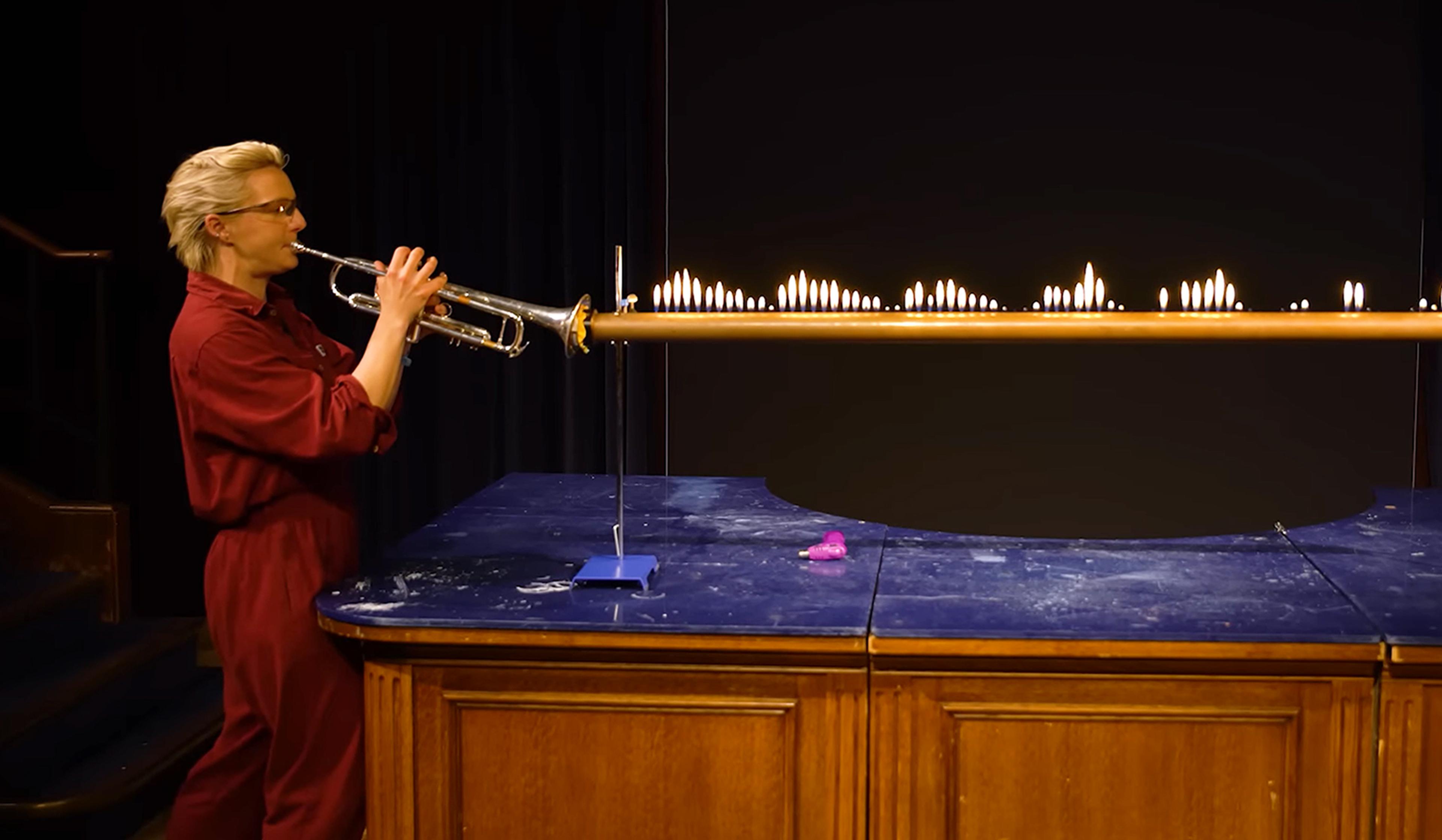 A person in a red jumpsuit playing the trumpet, causing flames along a metal tube to create visible sound waves.