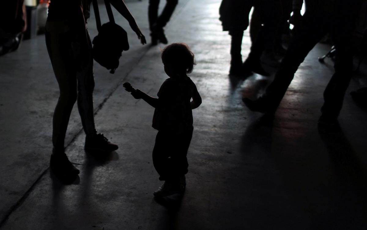 Silhouette of a child holding an object, surrounded by adults walking in a dimly lit environment.