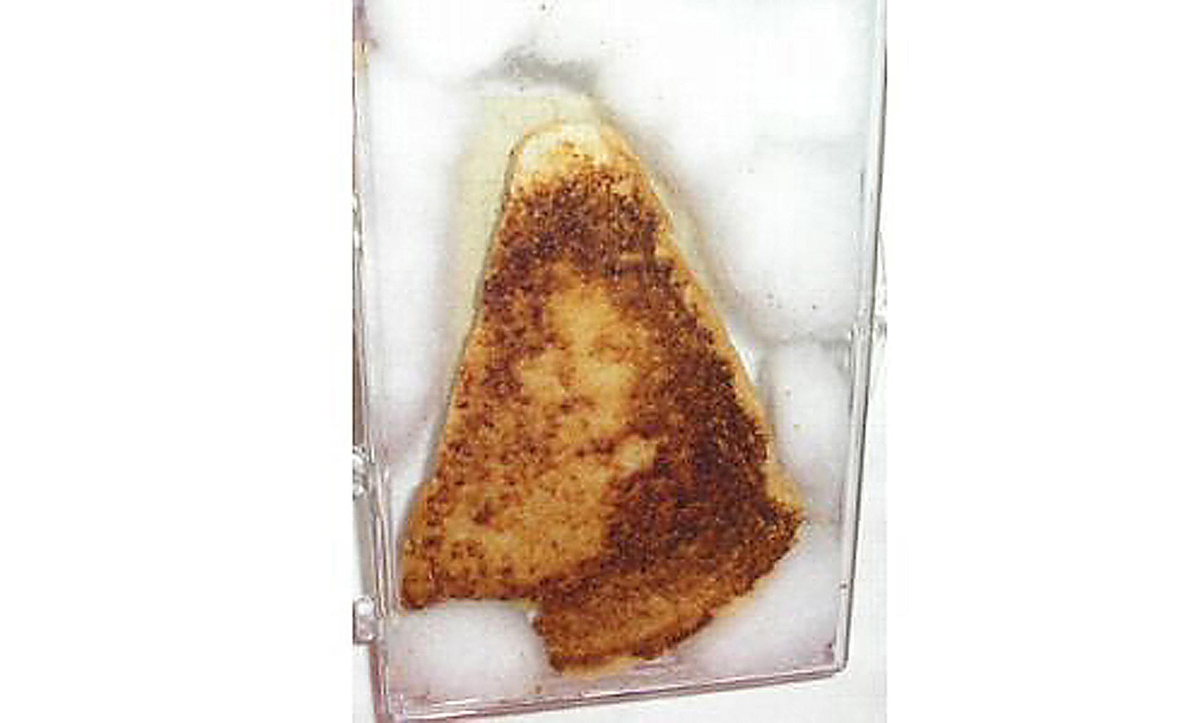 A slice of toasted bread with an indistinct image of a face on it, encased in a clear display case with white padding around it.