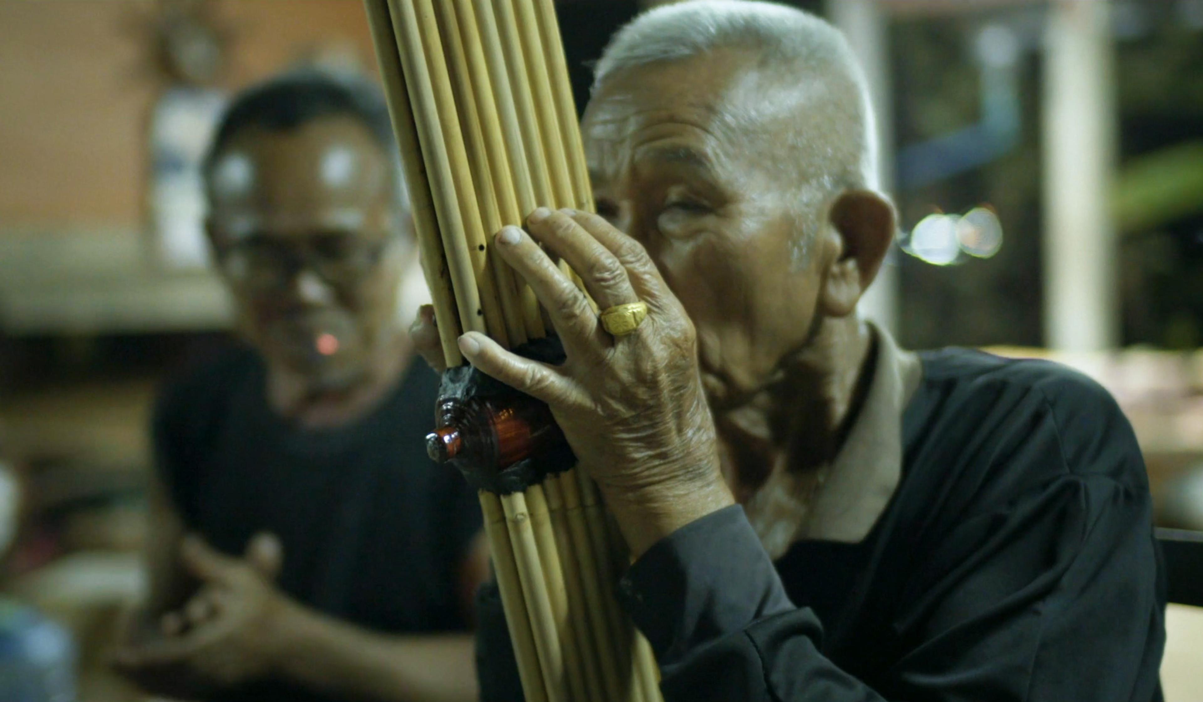 Elderly man playing a traditional bamboo musical instrument with another man in the background.