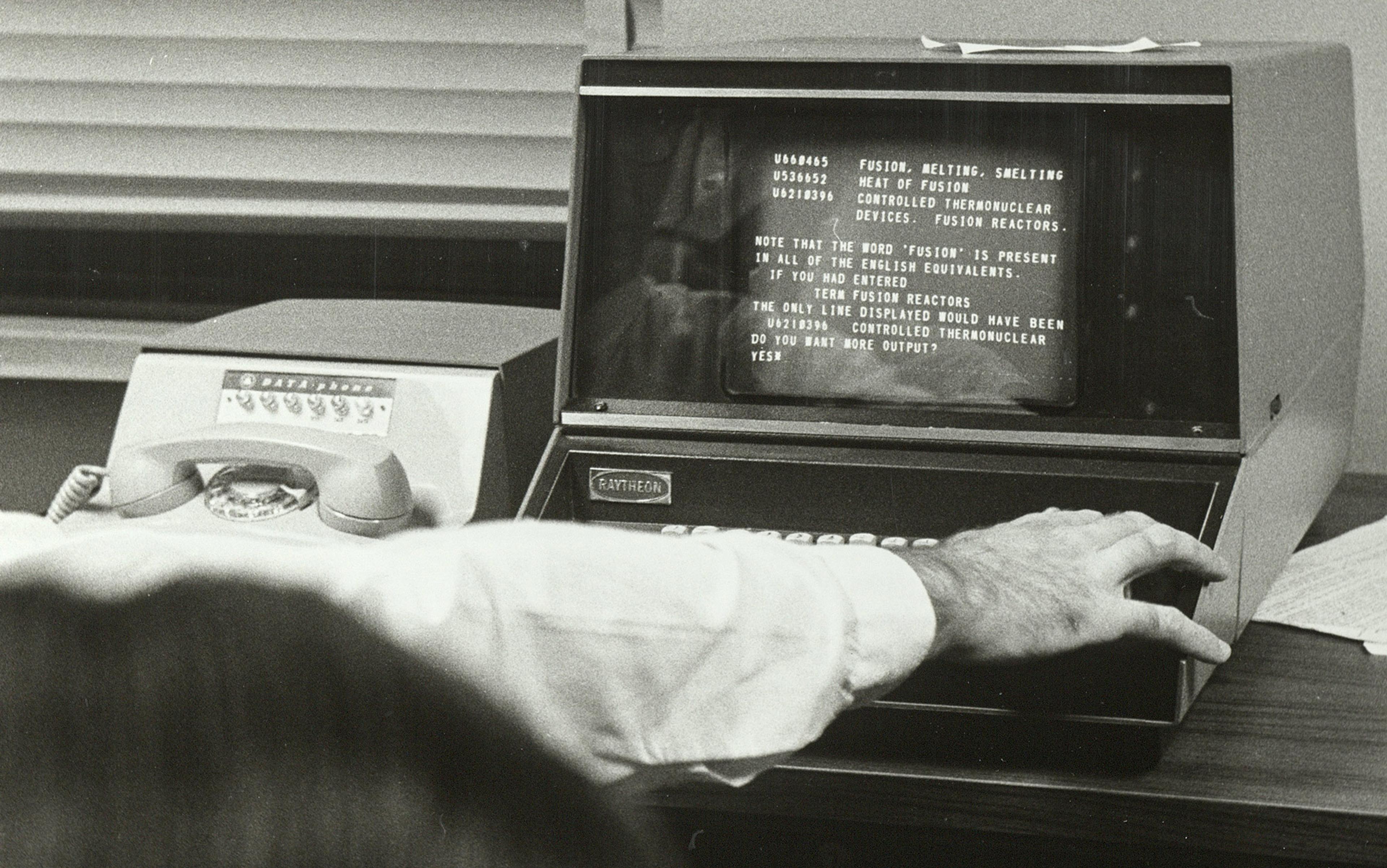 Person using a vintage Raytheon computer terminal with text on the screen, next to a rotary phone on a desk.