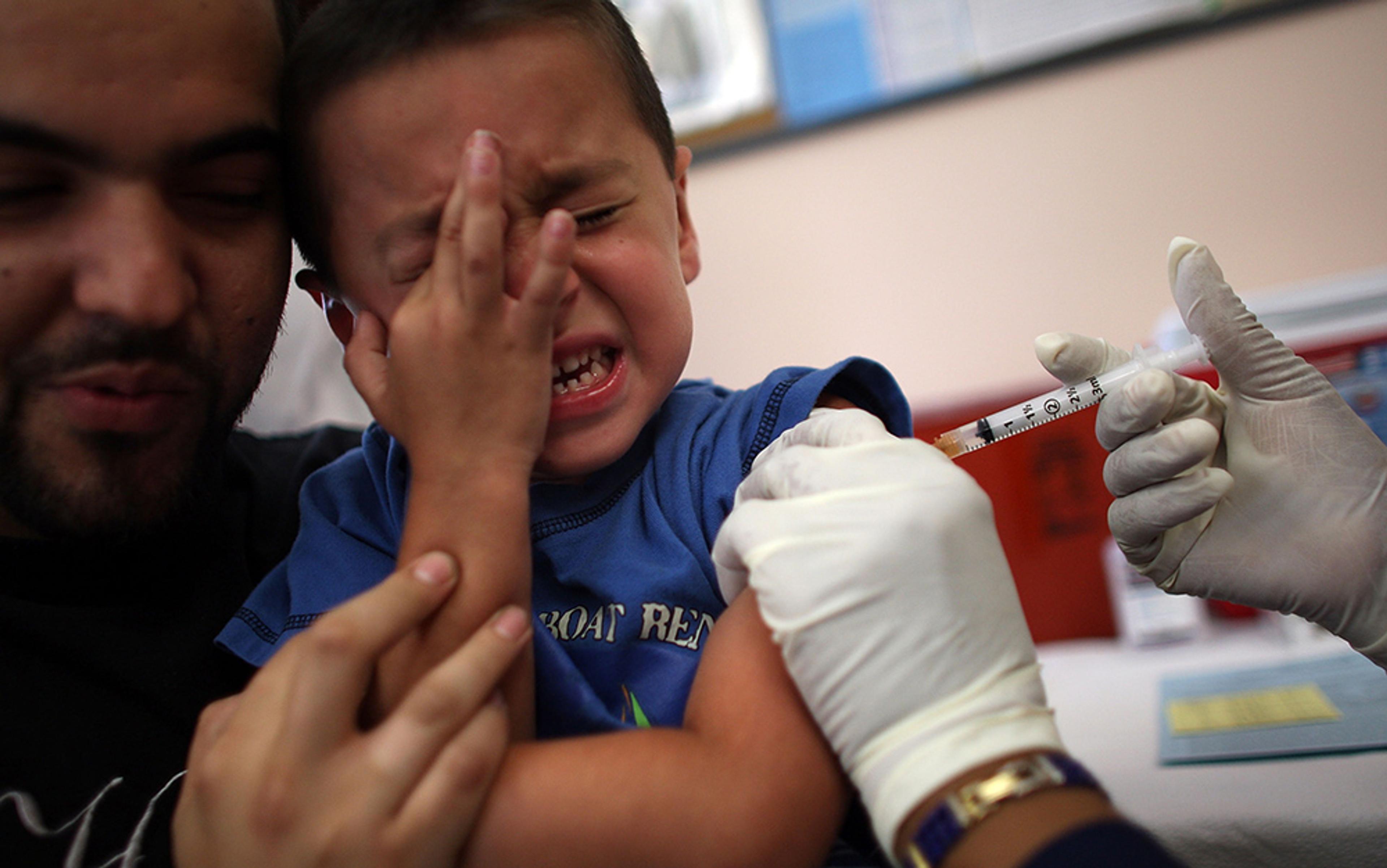 A young boy crying, eyes closed in pain, being held by an adult while receiving an injection from a gloved healthcare worker.