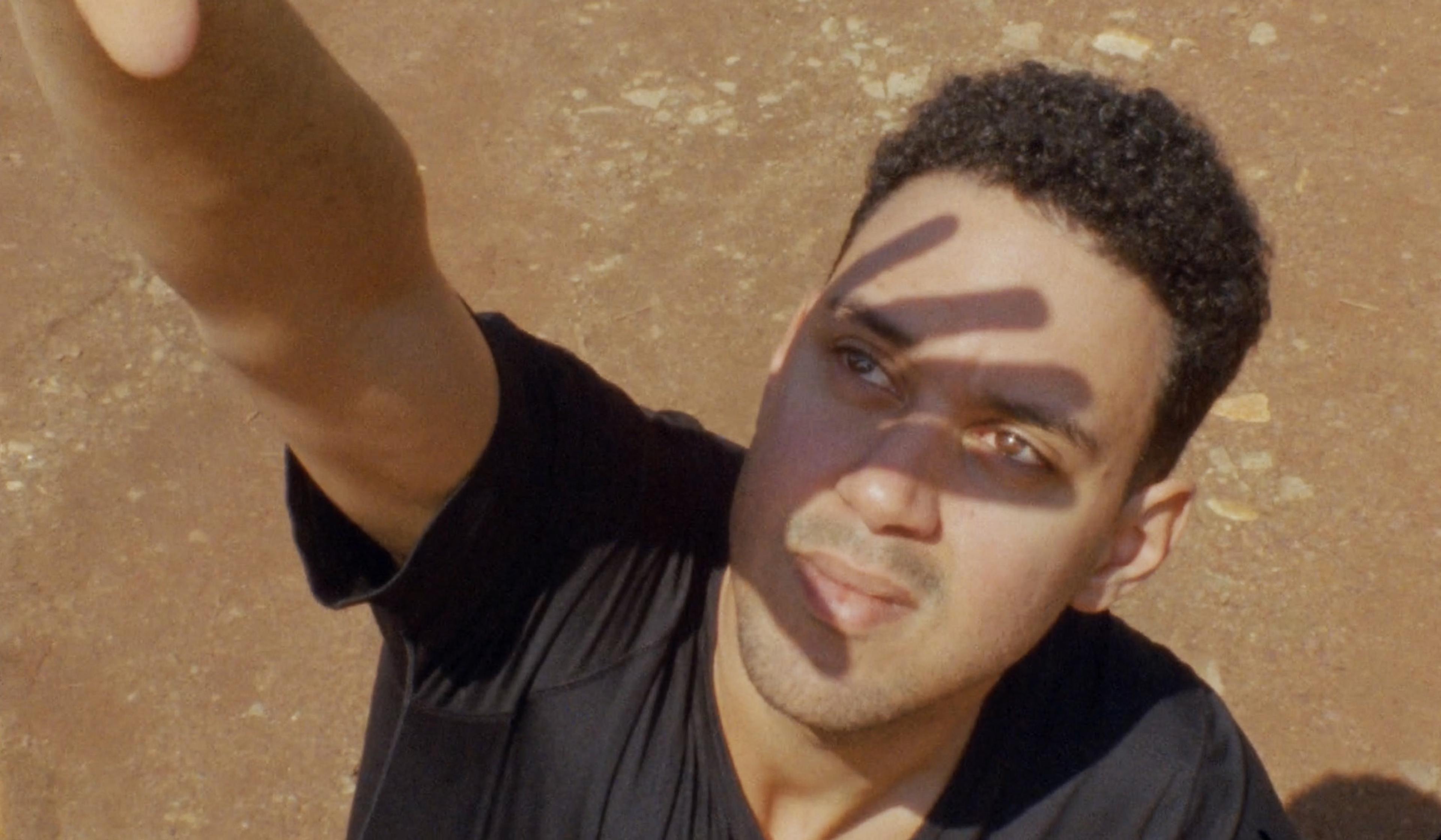 Man looking up while shielding his face from the sun with his hand, casting shadows on his face, in an outdoor setting with a dirt ground.