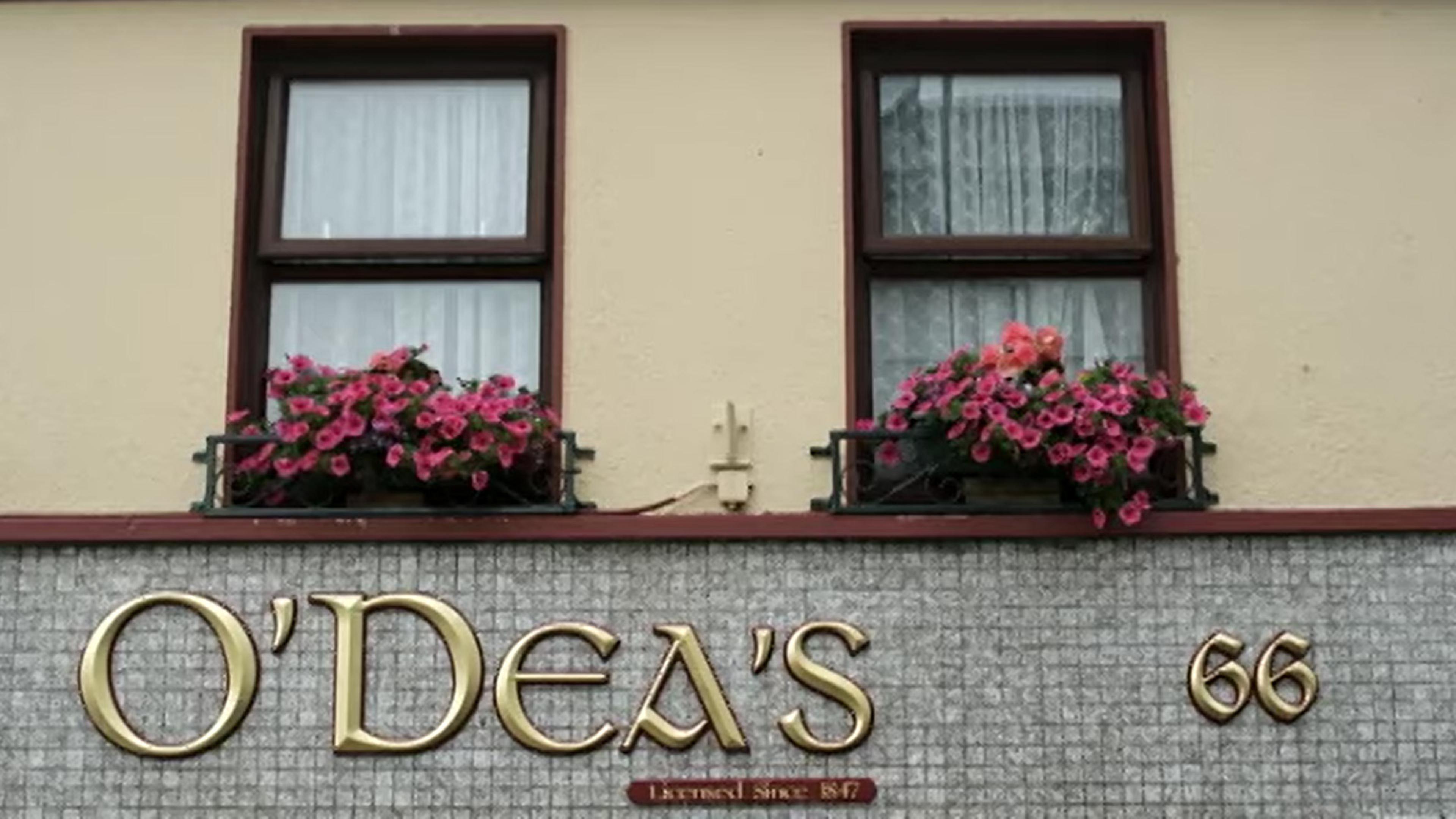 A shopfront with “O’DEA’S” in gold letters, two windows above with flower boxes holding pink flowers, and the number 66 on the right side.