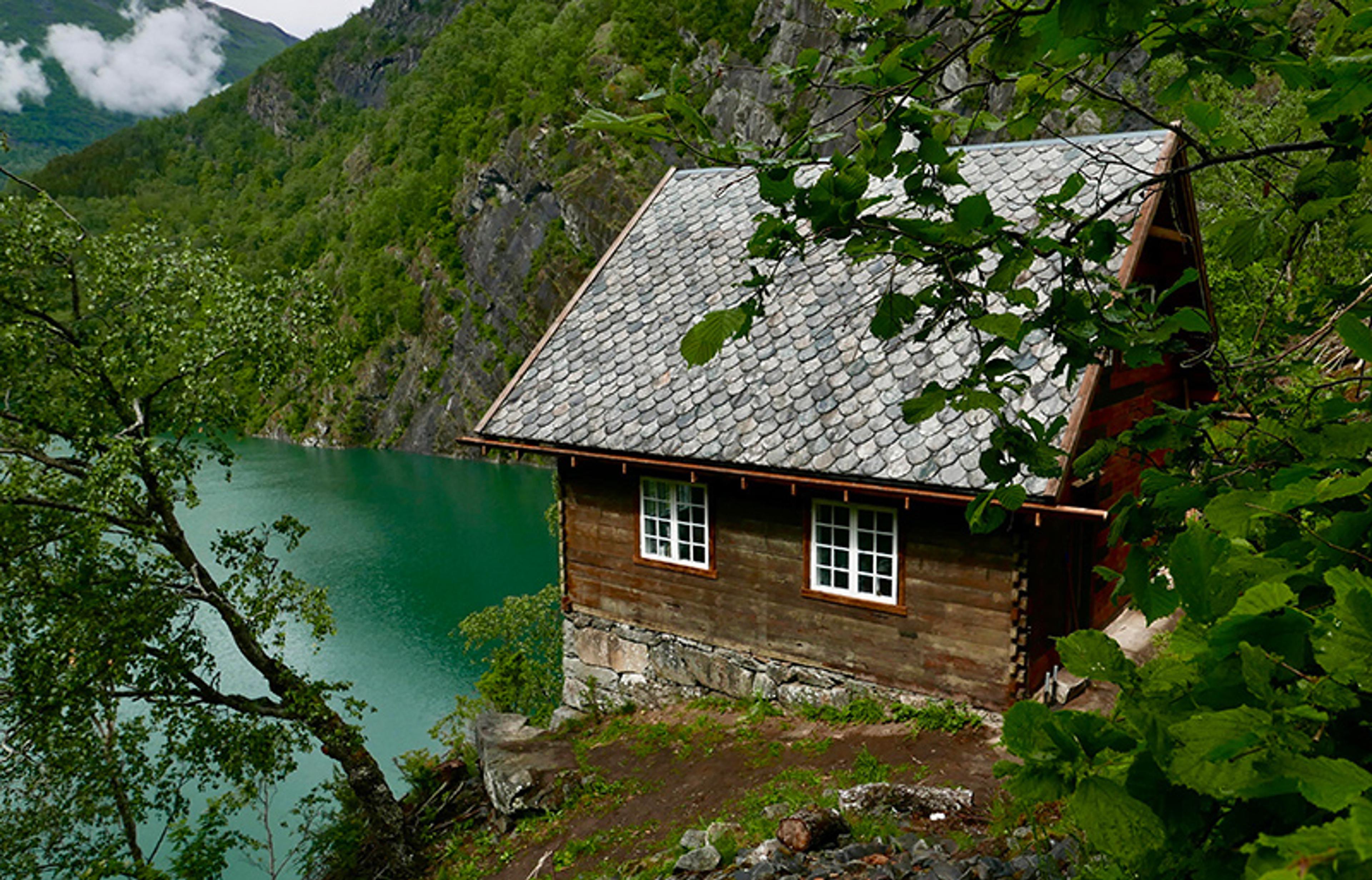 A rustic wooden cabin with a tiled roof by a turquoise lake, surrounded by lush green mountains and trees.