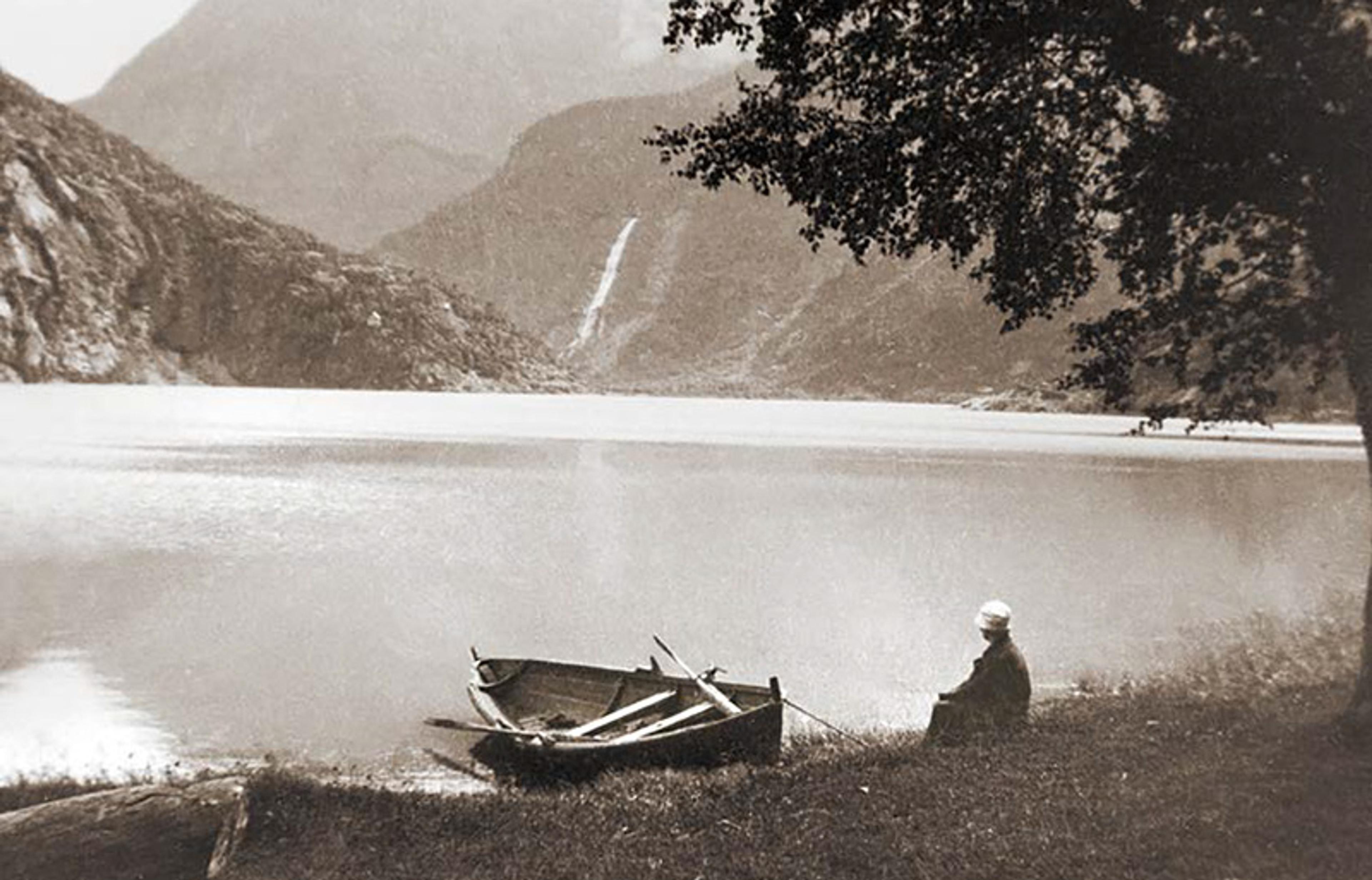 Vintage photo of a person sitting by a lake with a rowboat, surrounded by mountains and trees, watching a distant waterfall.