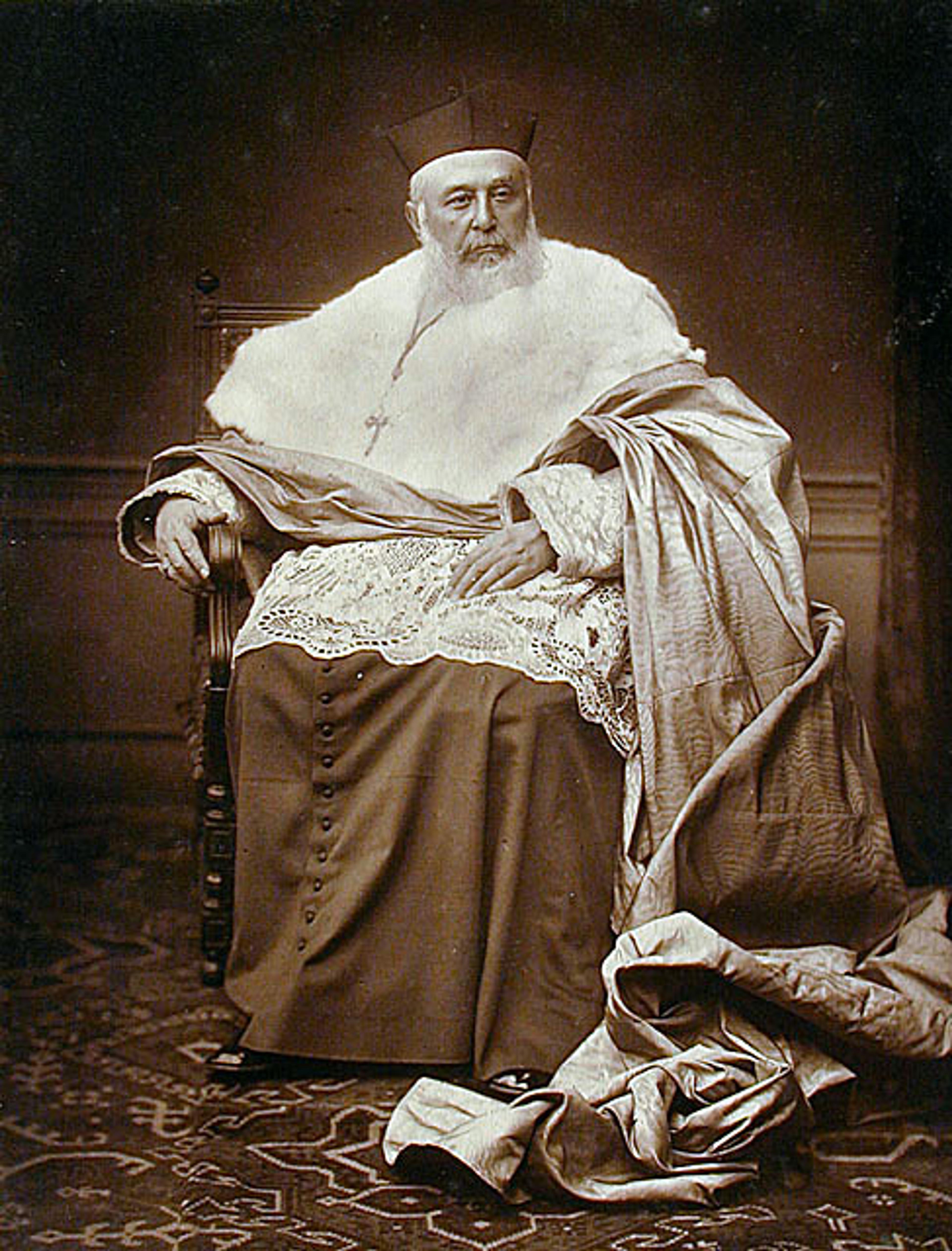 A sepia-toned photograph of an elderly man in ornate religious attire sitting on a chair with a serious expression.