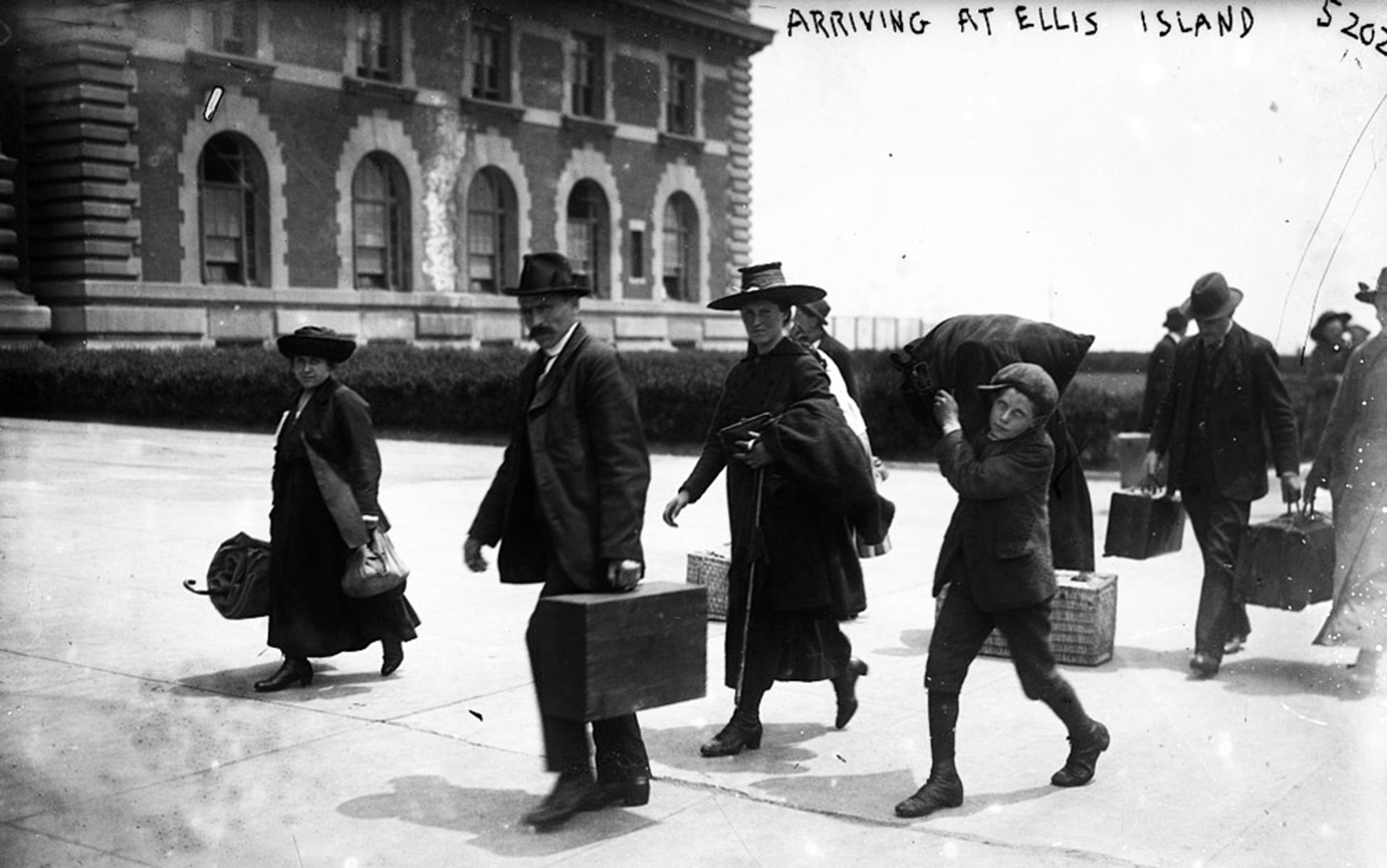 Vintage photo of people carrying luggage, labelled “Arriving at Ellis Island,” with a brick building in the background.