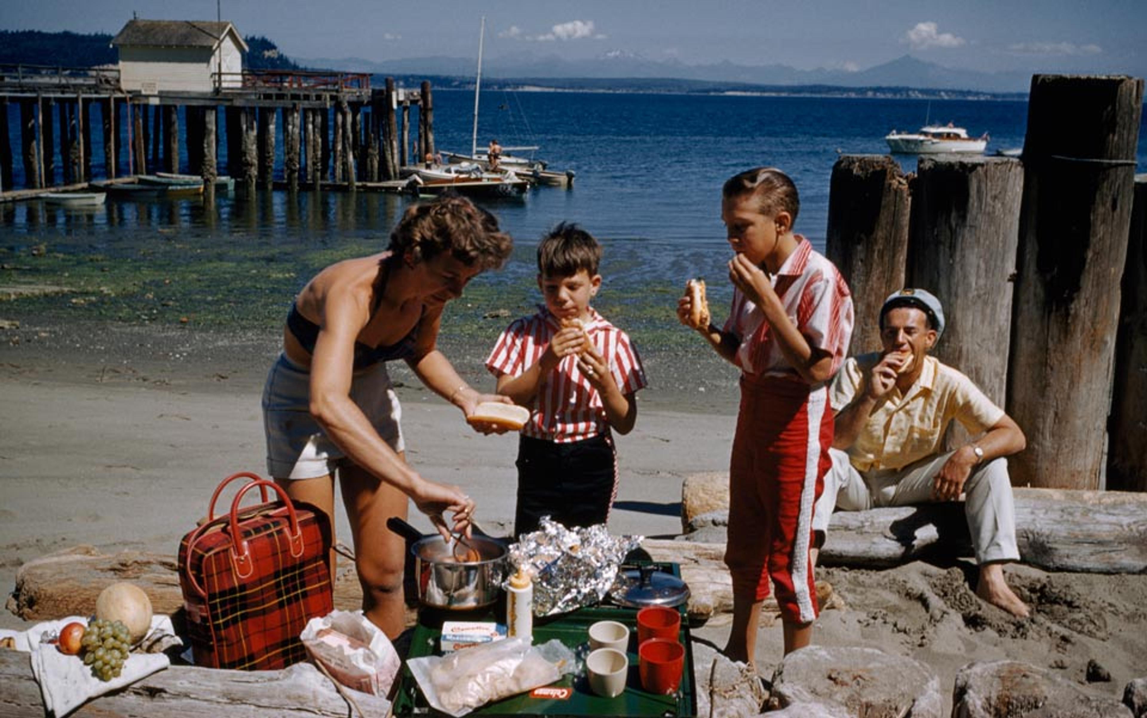 A family having a picnic on a beach with a pier in the background and a boat in the sea in the distance.